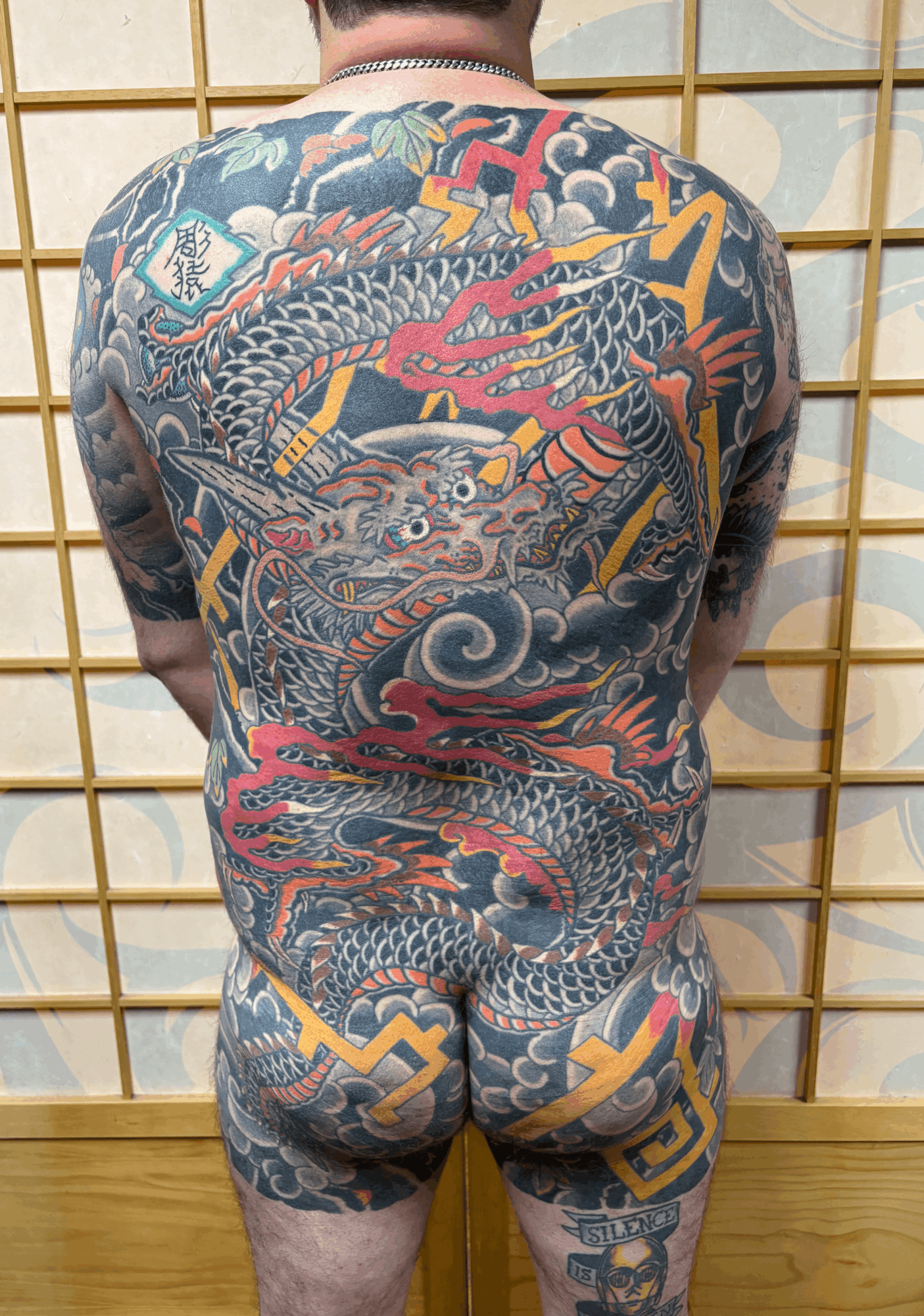 Back view of a person with a large, colorful dragon tattoo covering their back, shoulders, and legs, standing in front of a wooden grid background.