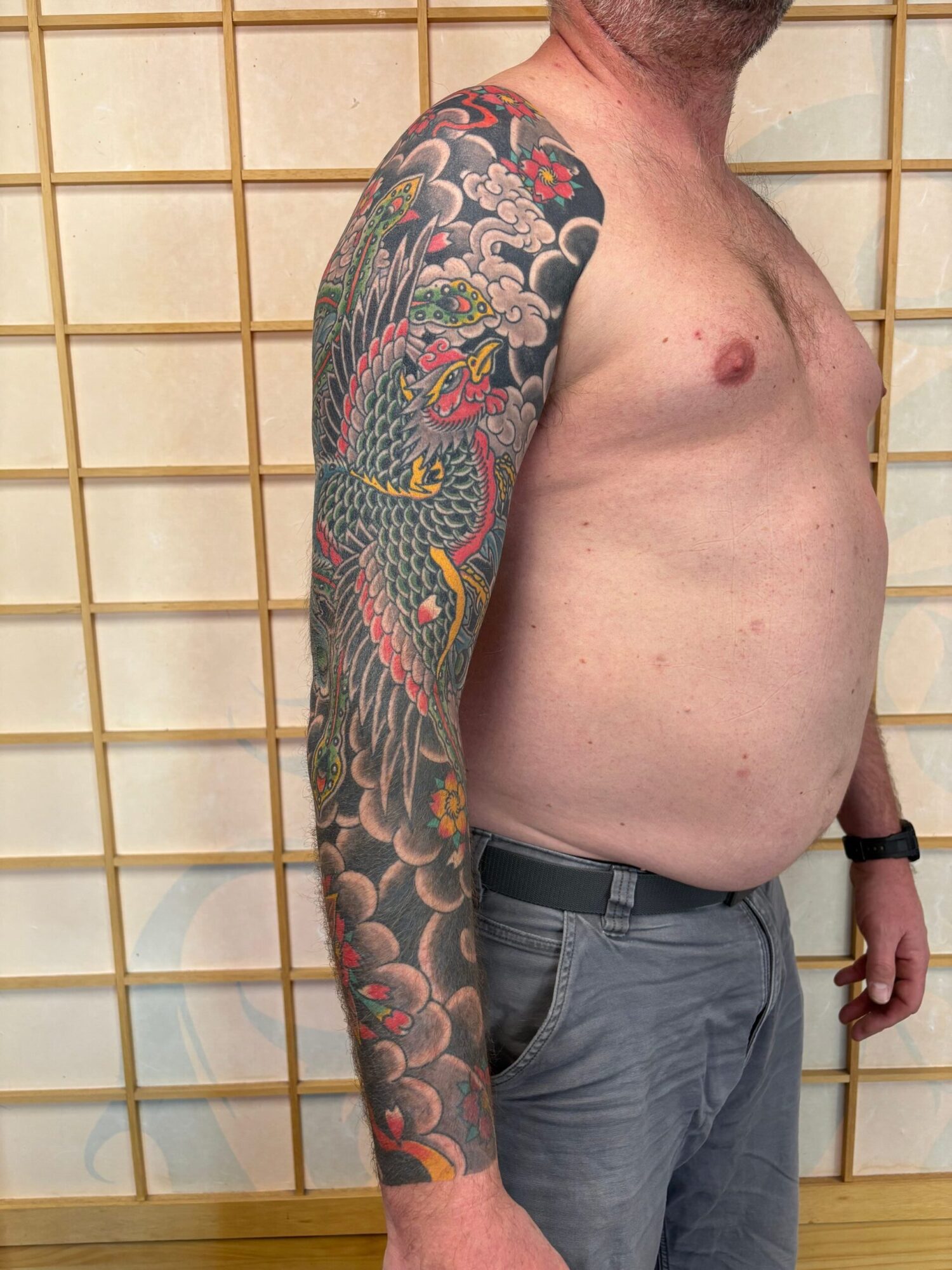 Person lying sideways with a colorful dragon tattoo on their arm, shirtless, against a wooden grid background.