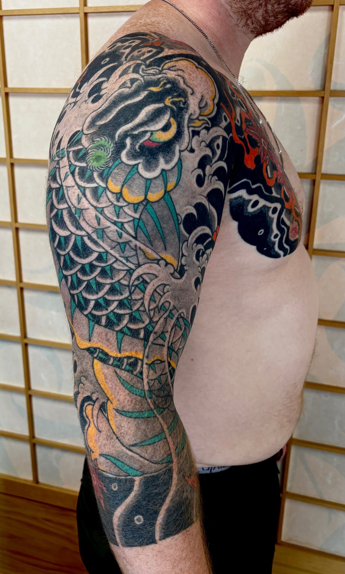 Tattooed arm and shoulder with colorful traditional Japanese-style dragon design, detailed scales, clouds, and waves, on a person's upper body.