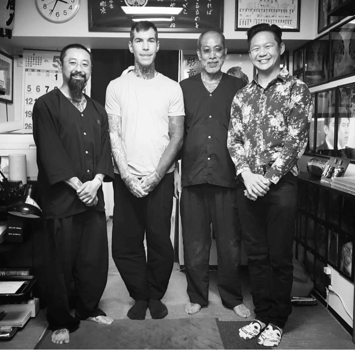 Four men standing indoors, smiling, with shelves and a clock in the background.