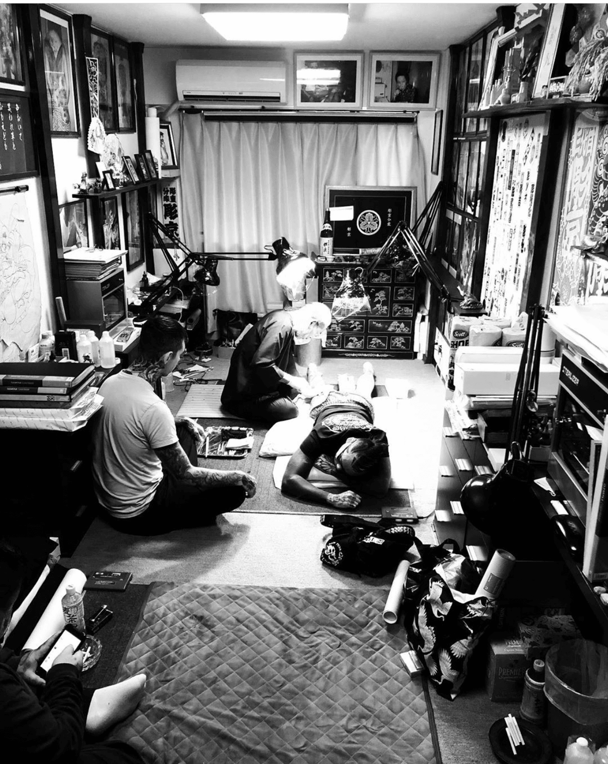 Four people sitting and kneeling on the floor in a room filled with bookshelves and various items, with a window and curtains in the background.