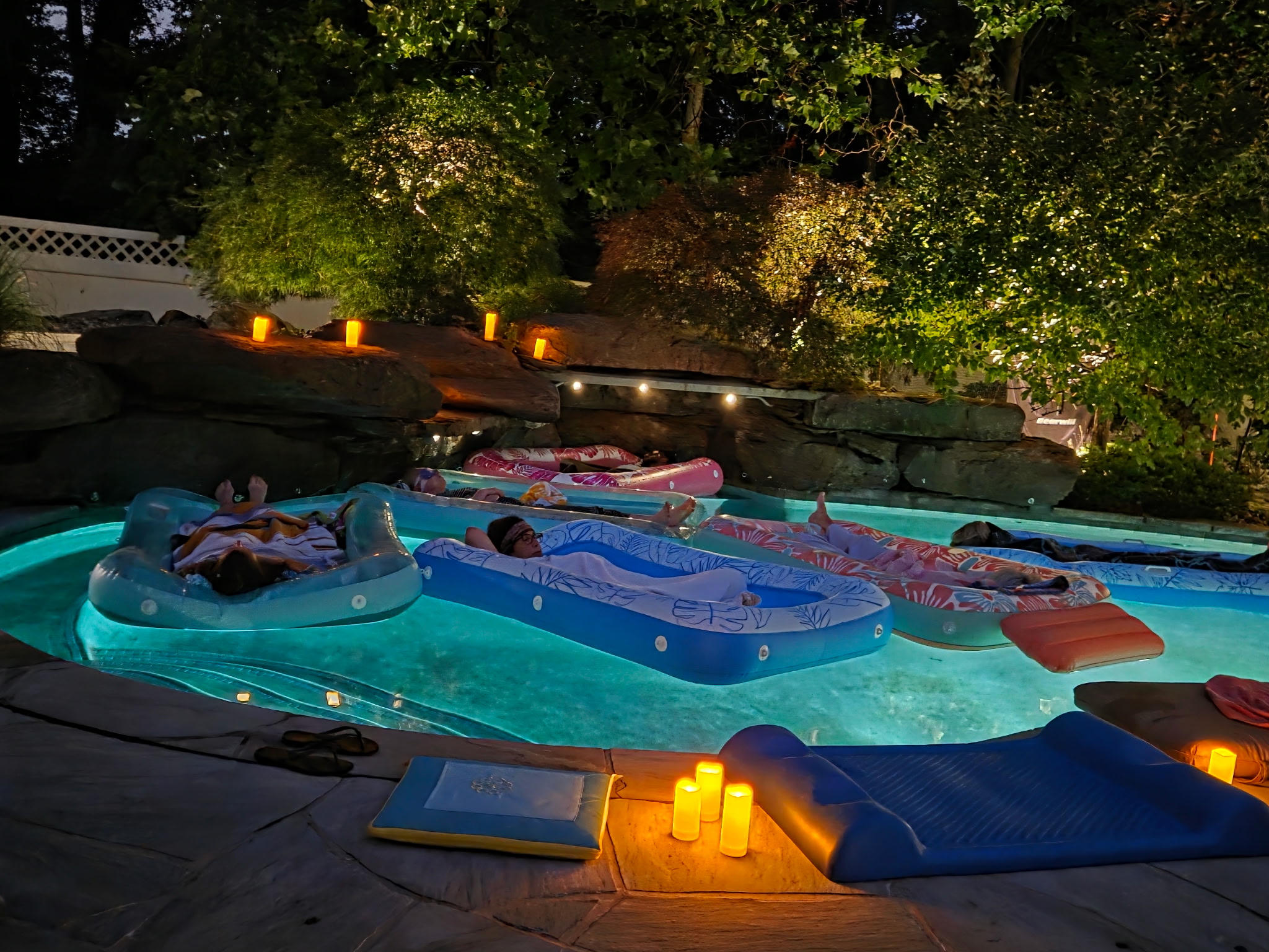 People relaxing on inflatable floats in a swimming pool at night, surrounded by trees and lit candles.