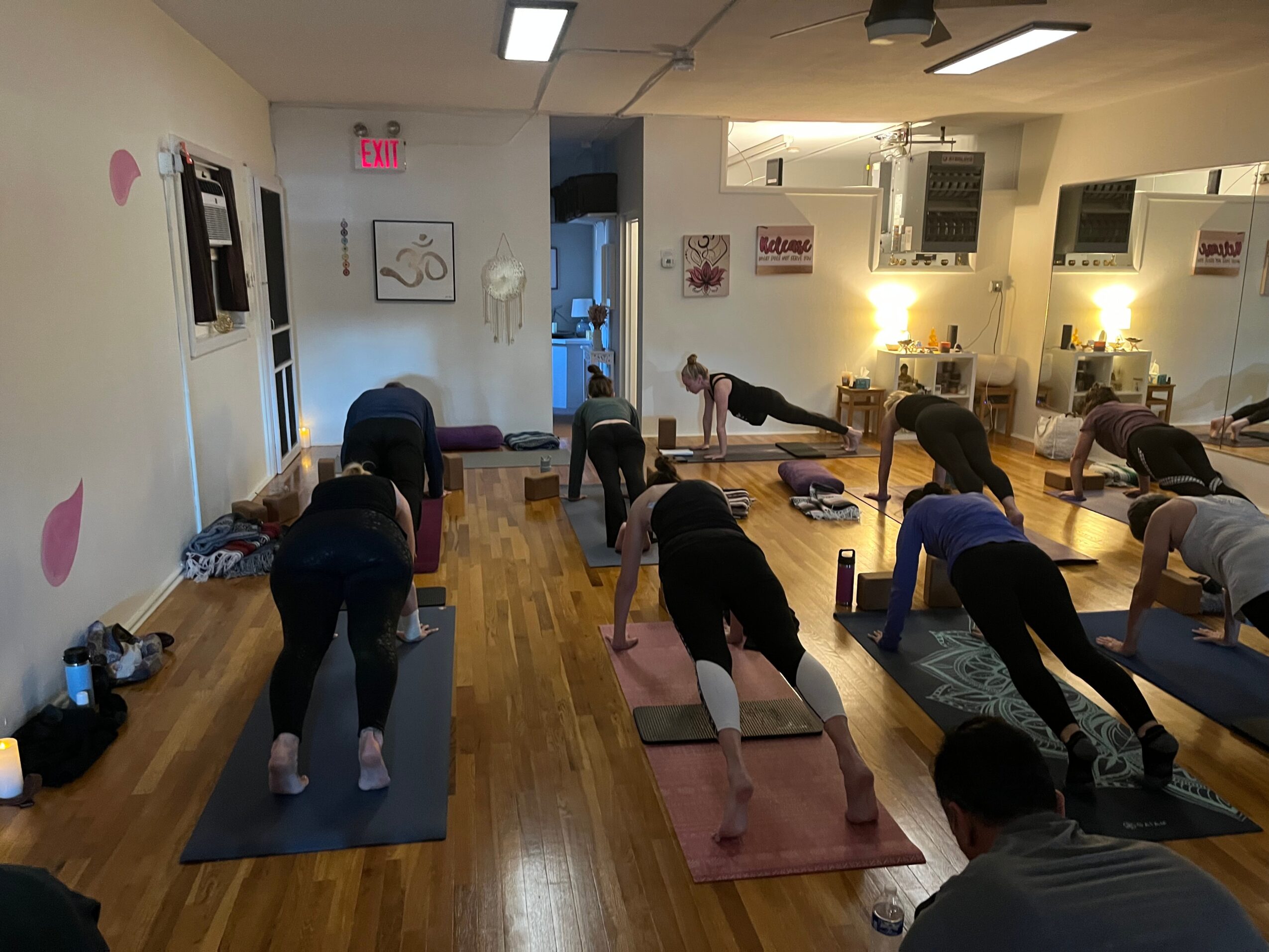 Group of people practicing yoga in a studio with wooden floors, mats, and wall decorations, facing downward in a plank pose.