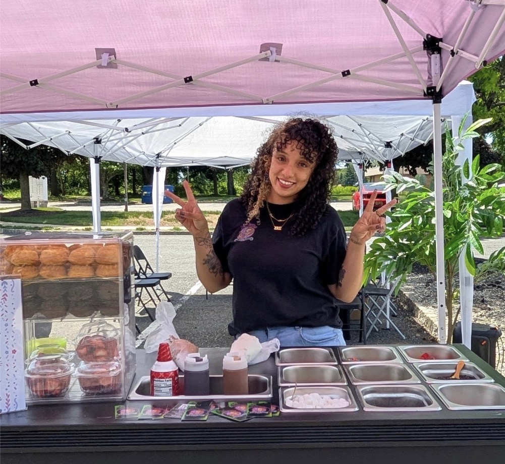 Young woman with curly hair smiling and making a peace sign at a food stand outdoors under a pink canopy, holding a red cup.