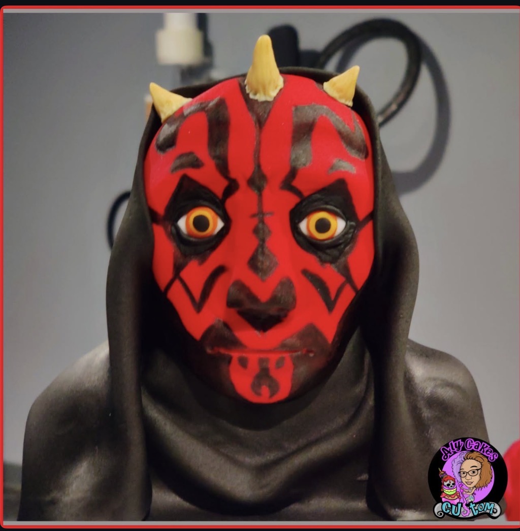 Person wearing a red and black mask with yellow horns and orange eyes, dressed in dark clothing, against a gray background.