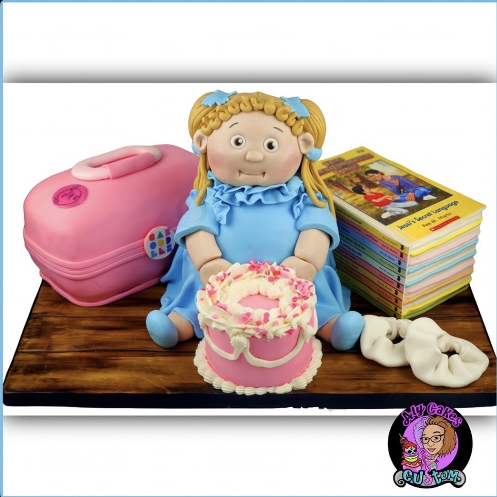 Doll with blonde hair, blue dress, sitting on a wooden surface, surrounded by a pink suitcase, books, a cake, and white objects.