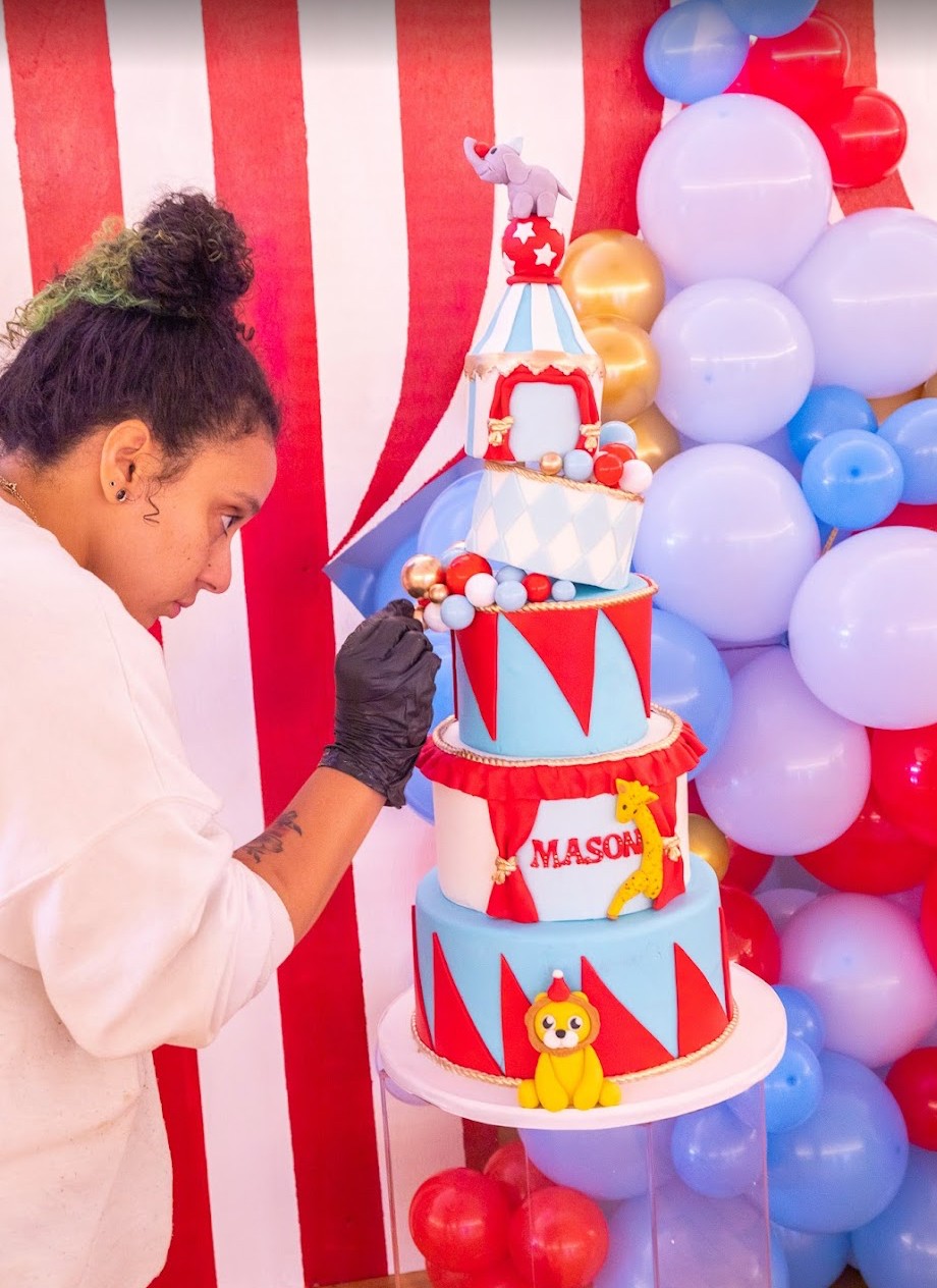 Girl decorating a circus-themed cake with balloons and striped background, wearing a white shirt and black glove.