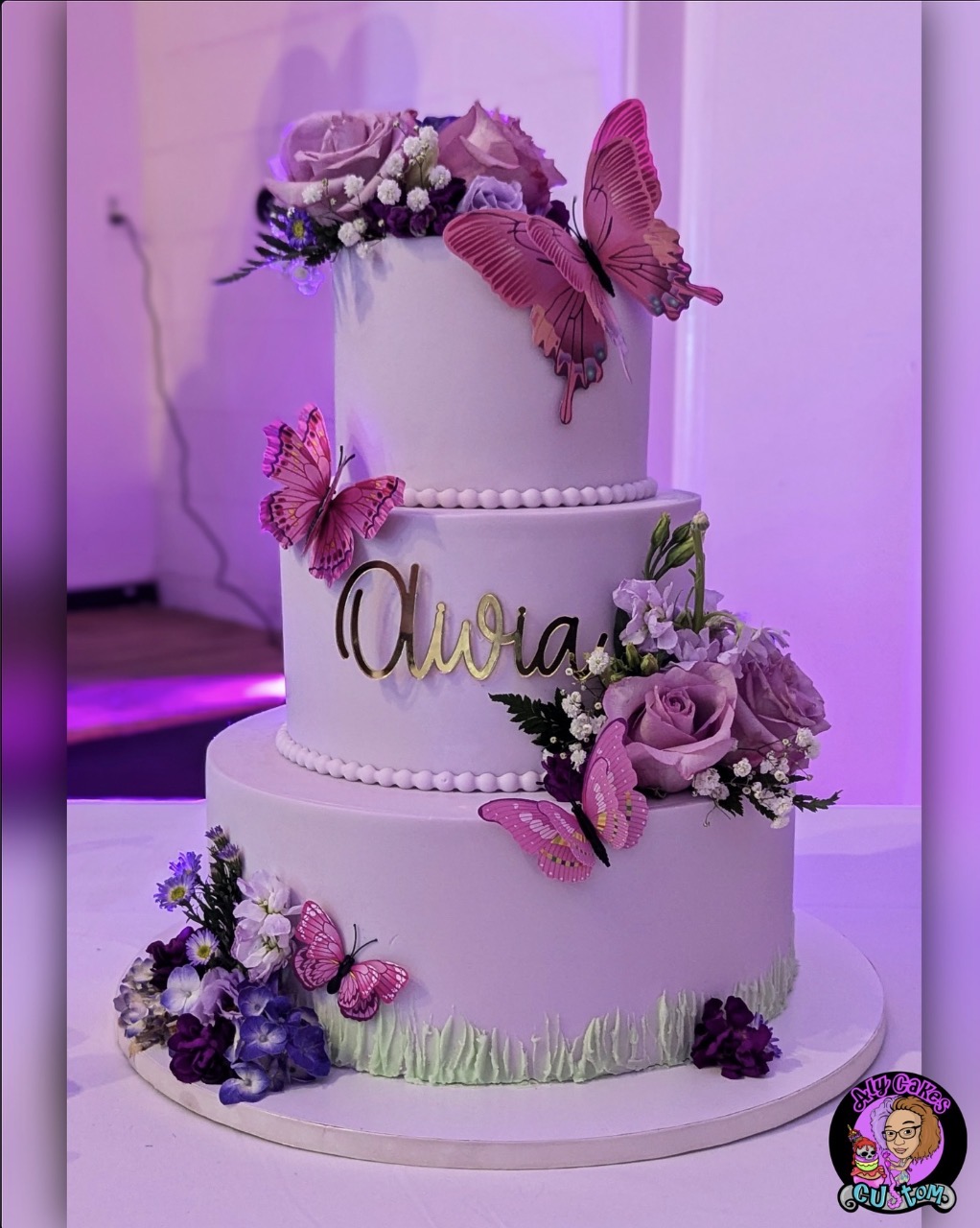Three-tier white cake decorated with pink and purple flowers, butterflies, and the name 'Olivia' in gold script.
