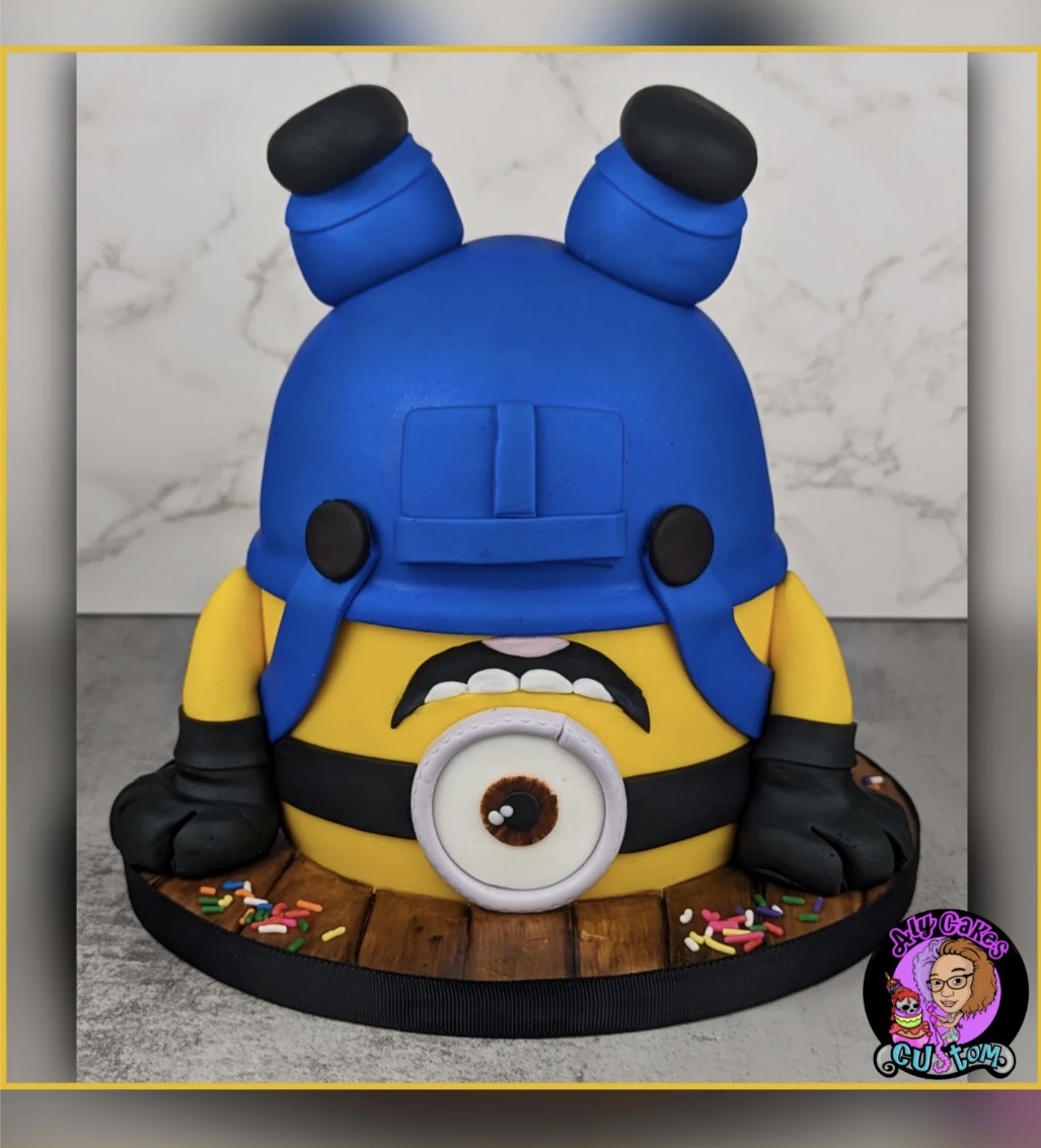 Cake shaped like a Minion character, upside down, with blue overalls, black gloves, and a large eye, on a black base.
