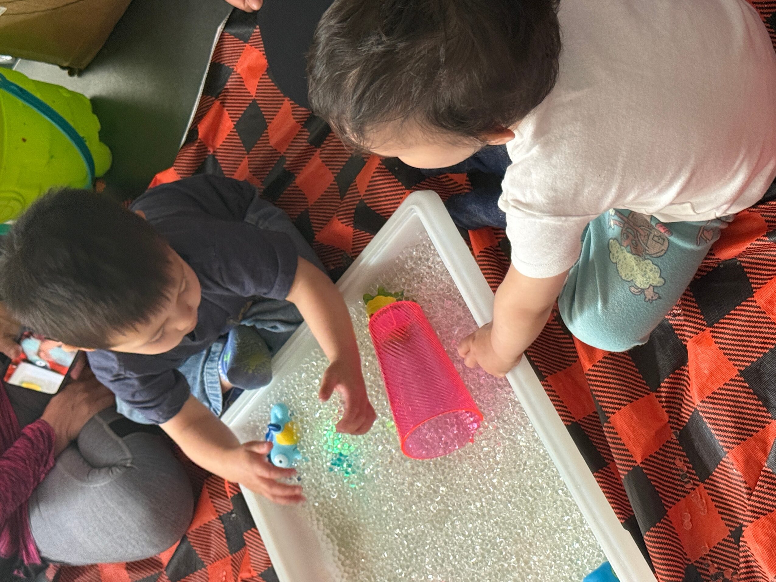 Two children playing with small colorful objects on a textured surface, with a pink container nearby.