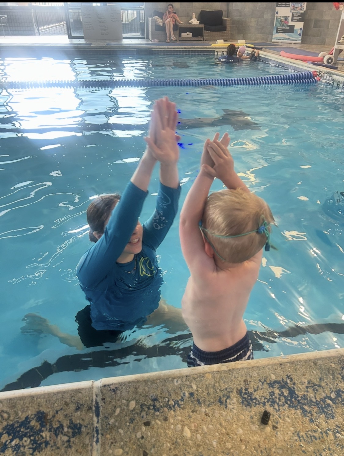 Two children in a swimming pool, facing each other, raising their hands for a high five, with other swimmers in the background.