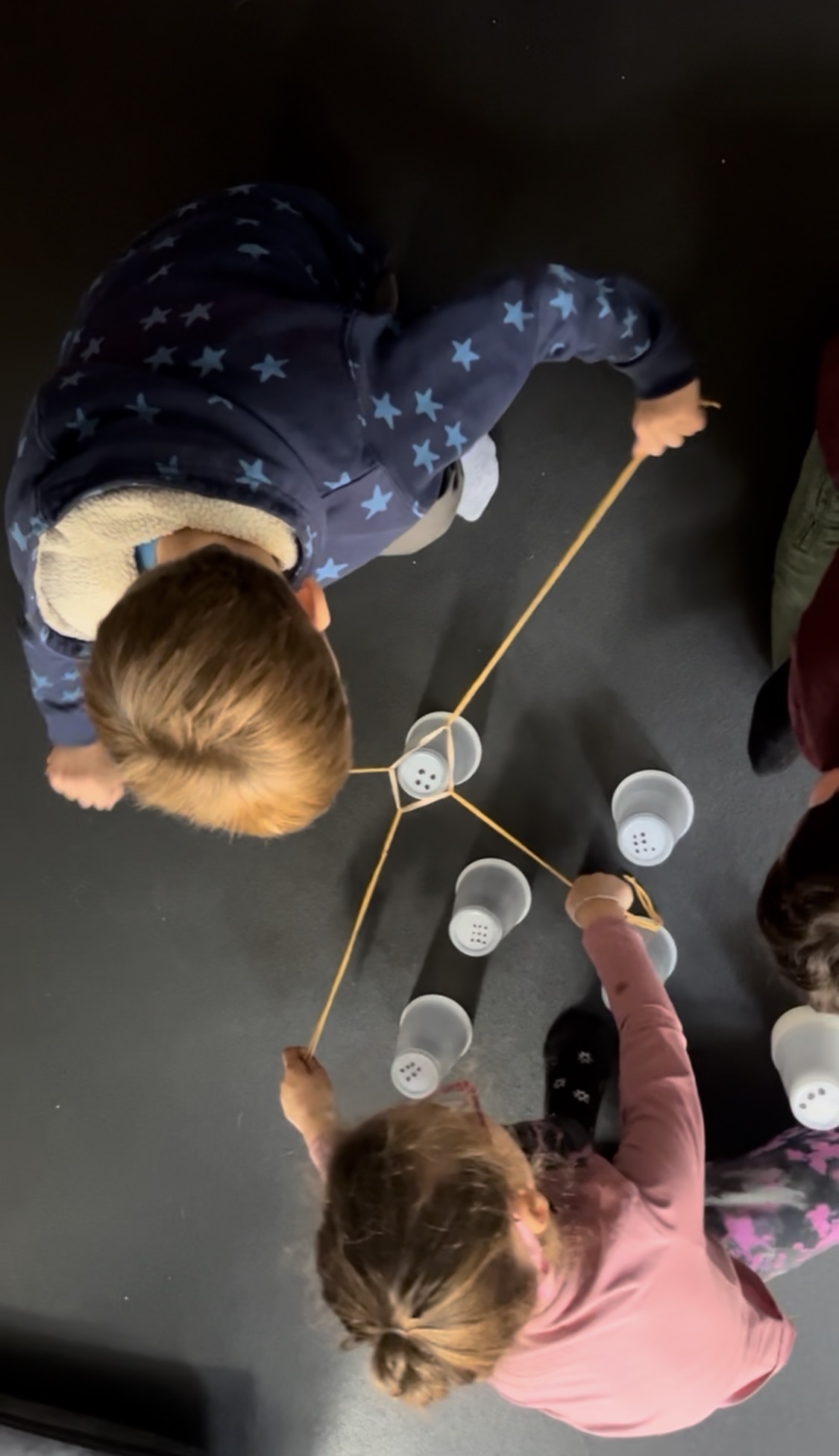 Two children playing with cups and strings on a dark surface, viewed from above.