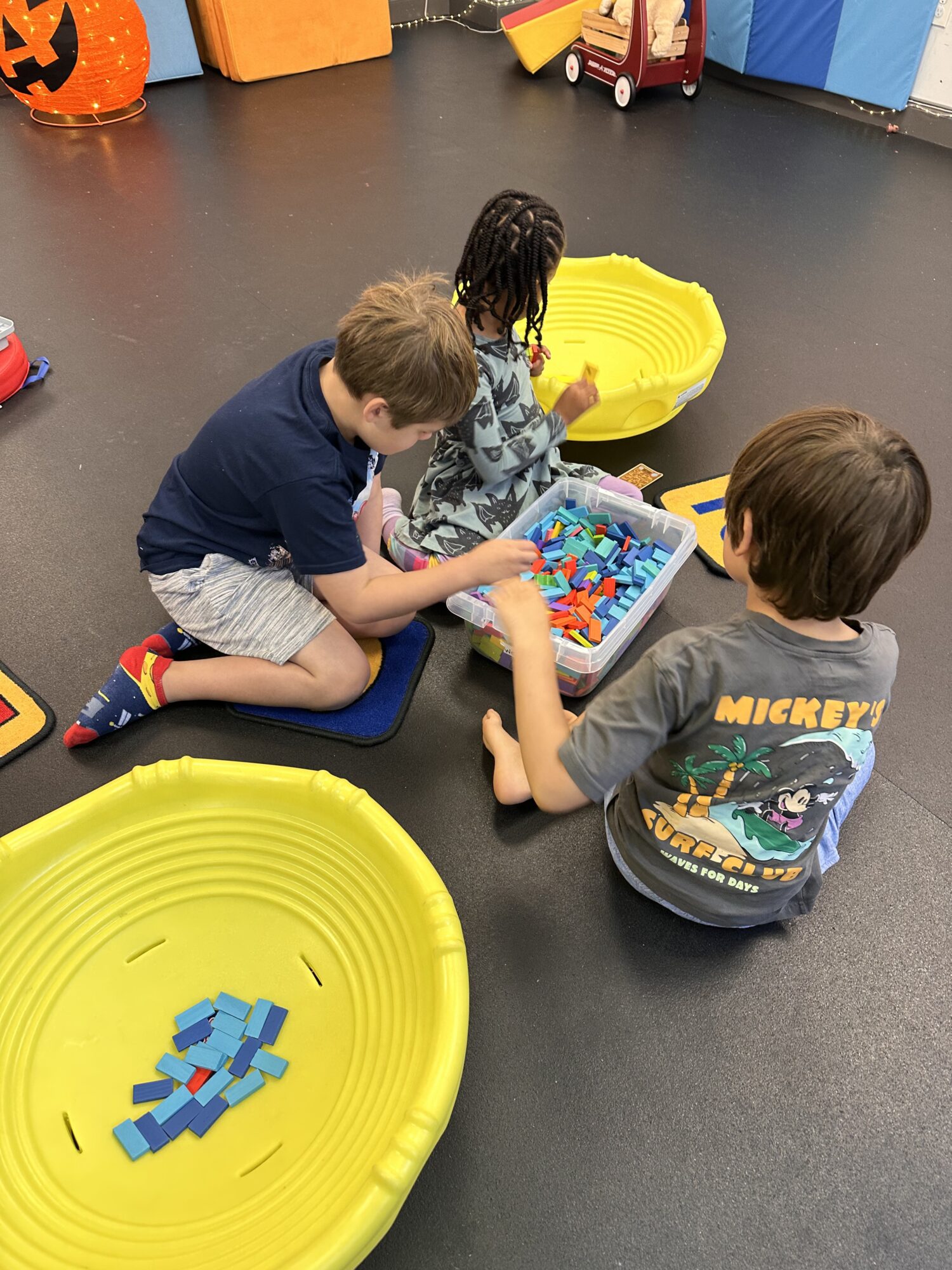 Three children sit on the floor playing with colorful building blocks in a playroom, with yellow containers nearby.