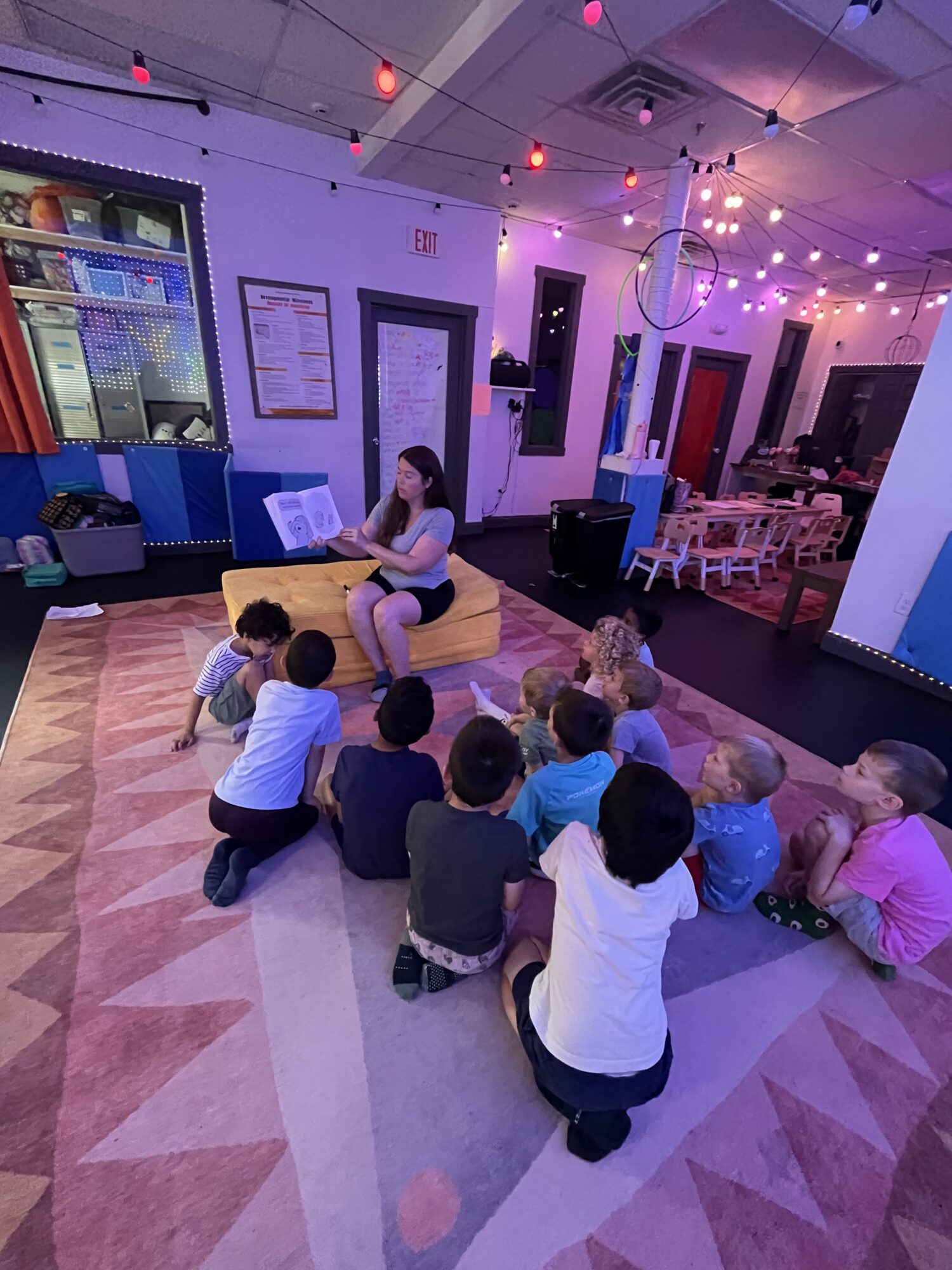 A woman sits on a yellow chair reading to a group of children seated on a patterned carpet in a colorful indoor space with string lights and decorations.