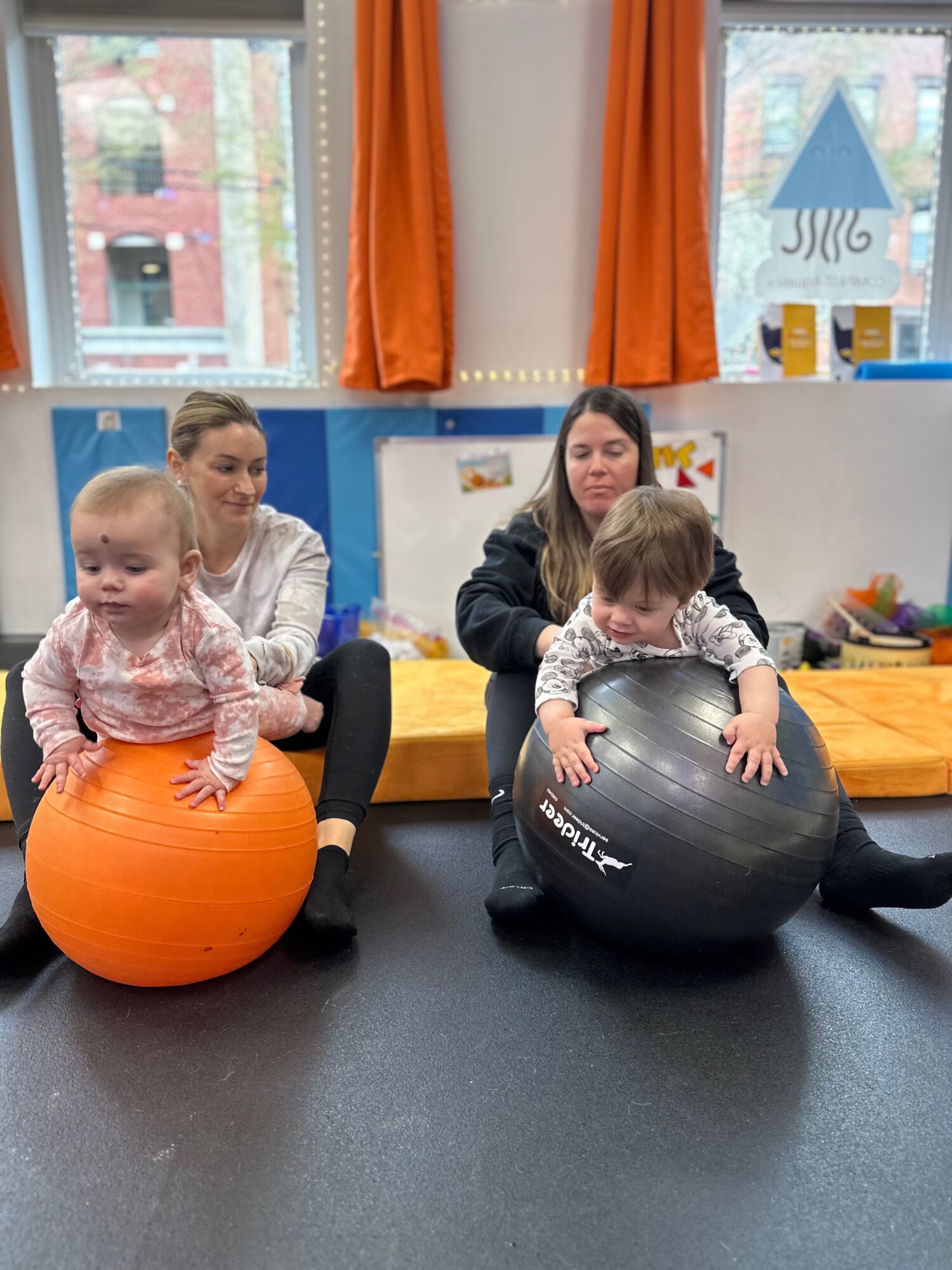 Two young children sit on large exercise balls, with two women in a room with windows and orange curtains.