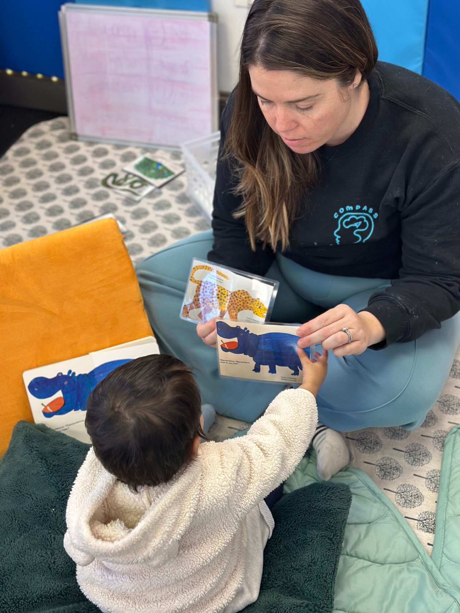 Adult woman reading a children's book to a young child sitting on the floor, with colorful cushions around them.