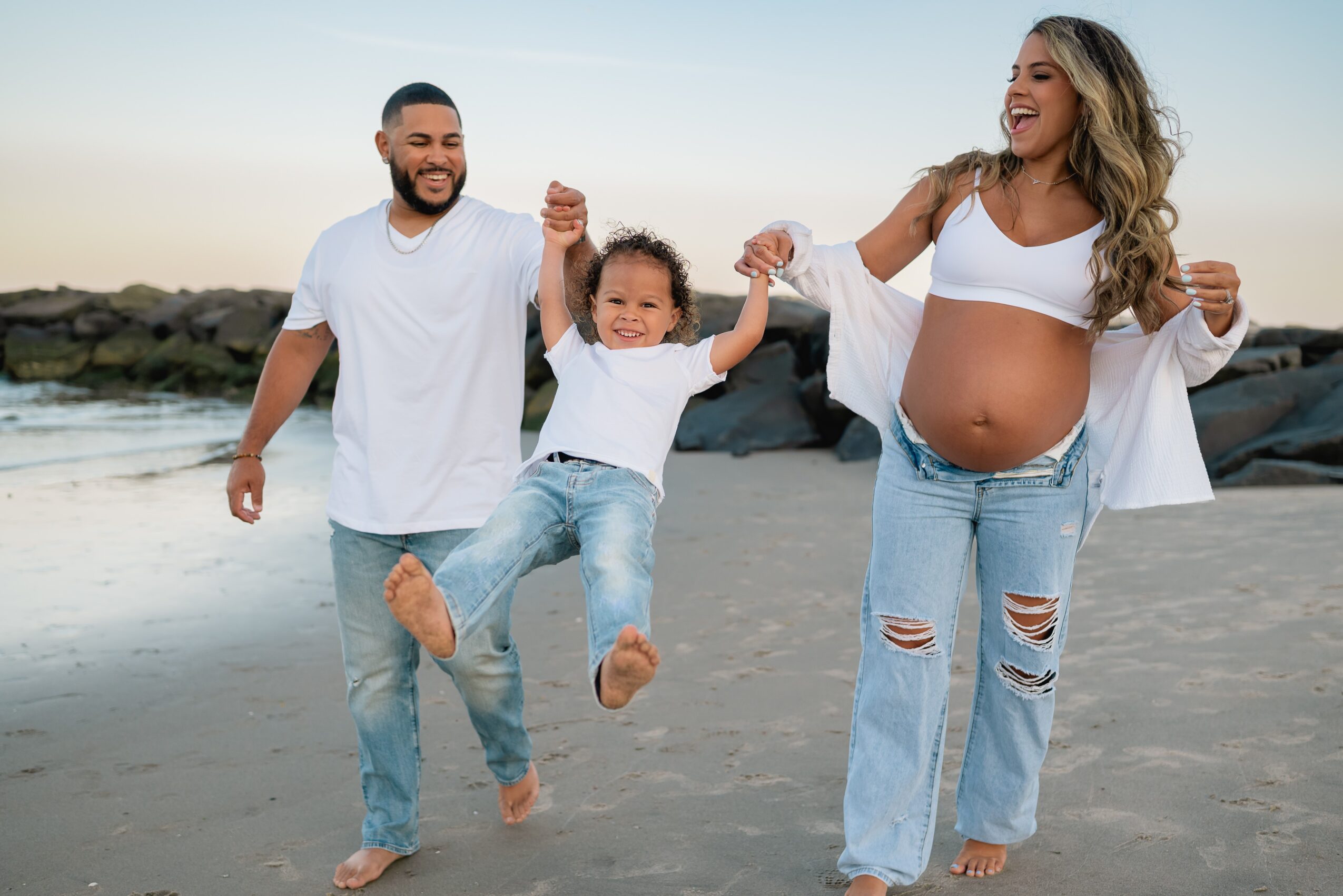 Family on the beach, a pregnant woman, a man, and a child holding hands and playing, smiling and enjoying the outdoors.