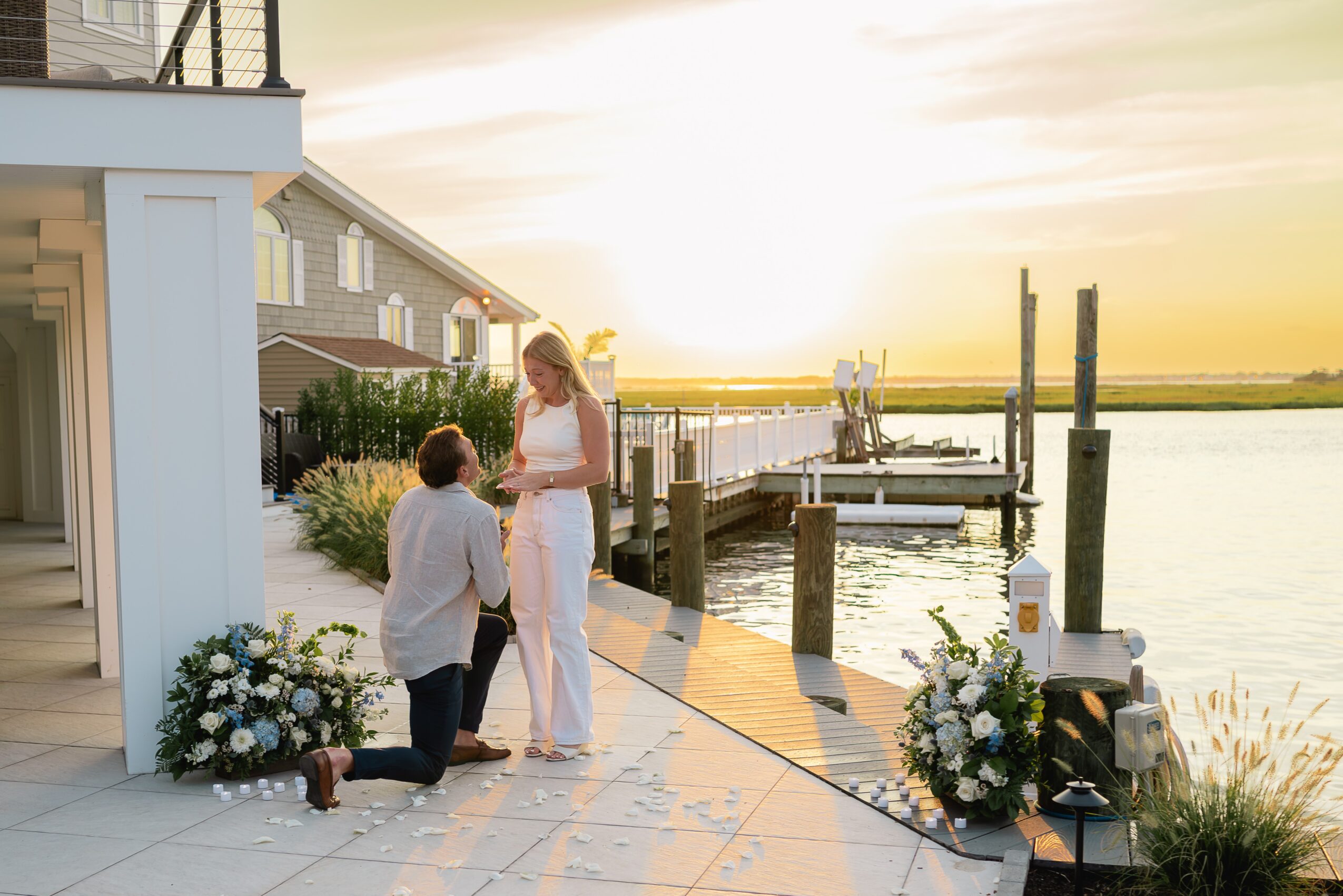 Person kneeling with a ring box, person standing, and a woman in white dress near water at sunset, with floral arrangements nearby.