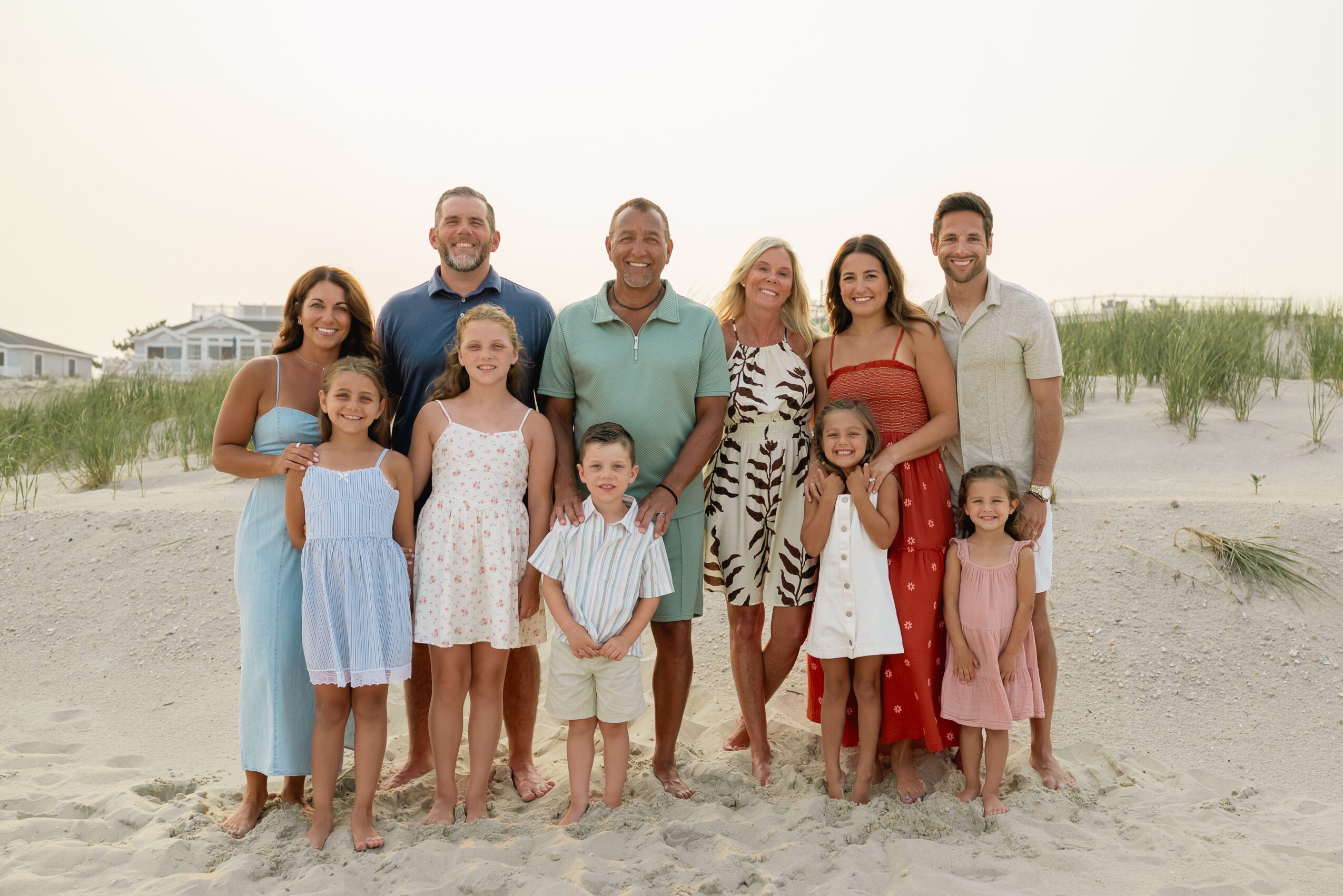 Group of ten people, including children and adults, standing on a sandy beach with grass and buildings in background.