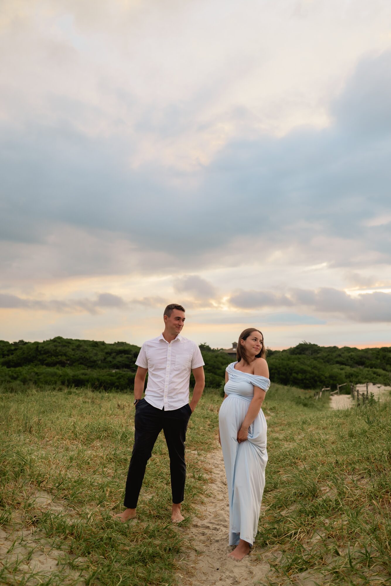 A man and a pregnant woman stand outdoors on a grassy field under a cloudy sky, with the woman holding her belly.