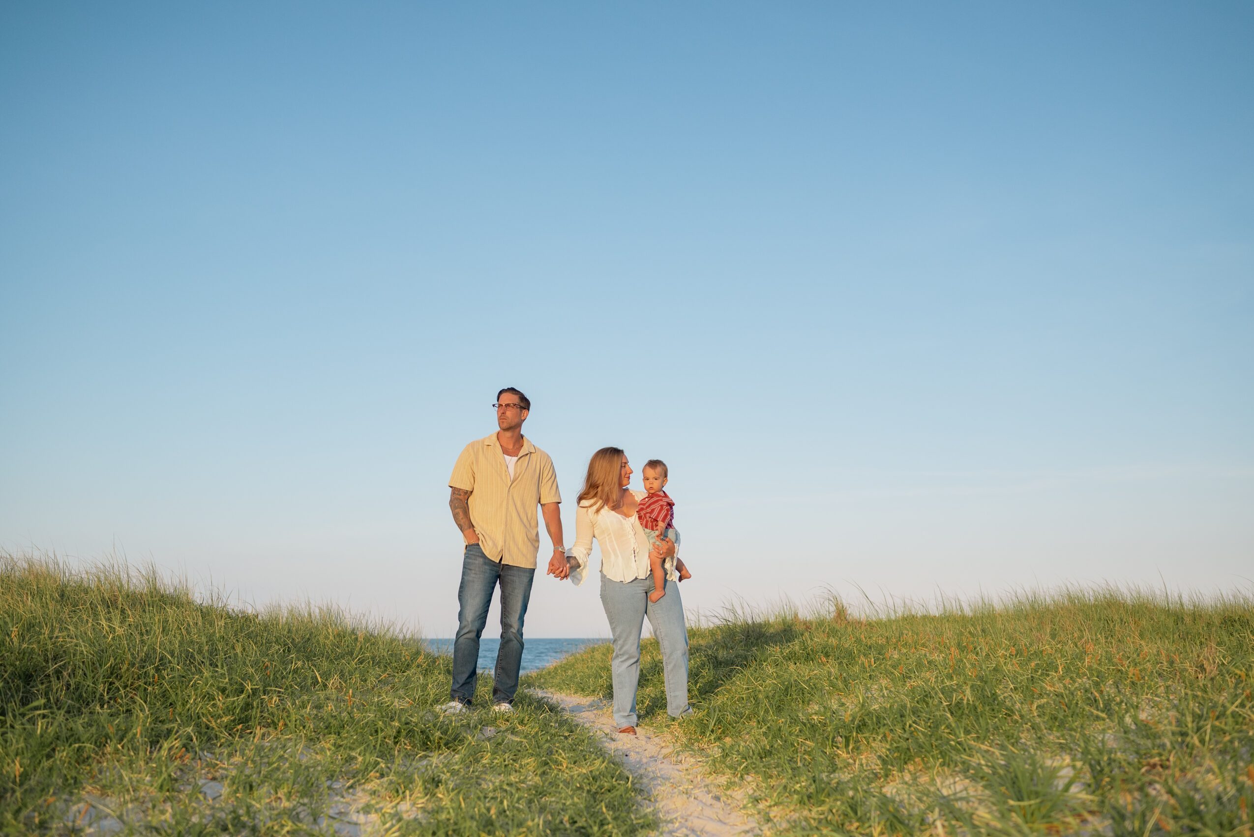 Family of three walking on a grassy hill under a clear blue sky, holding hands and carrying a child.