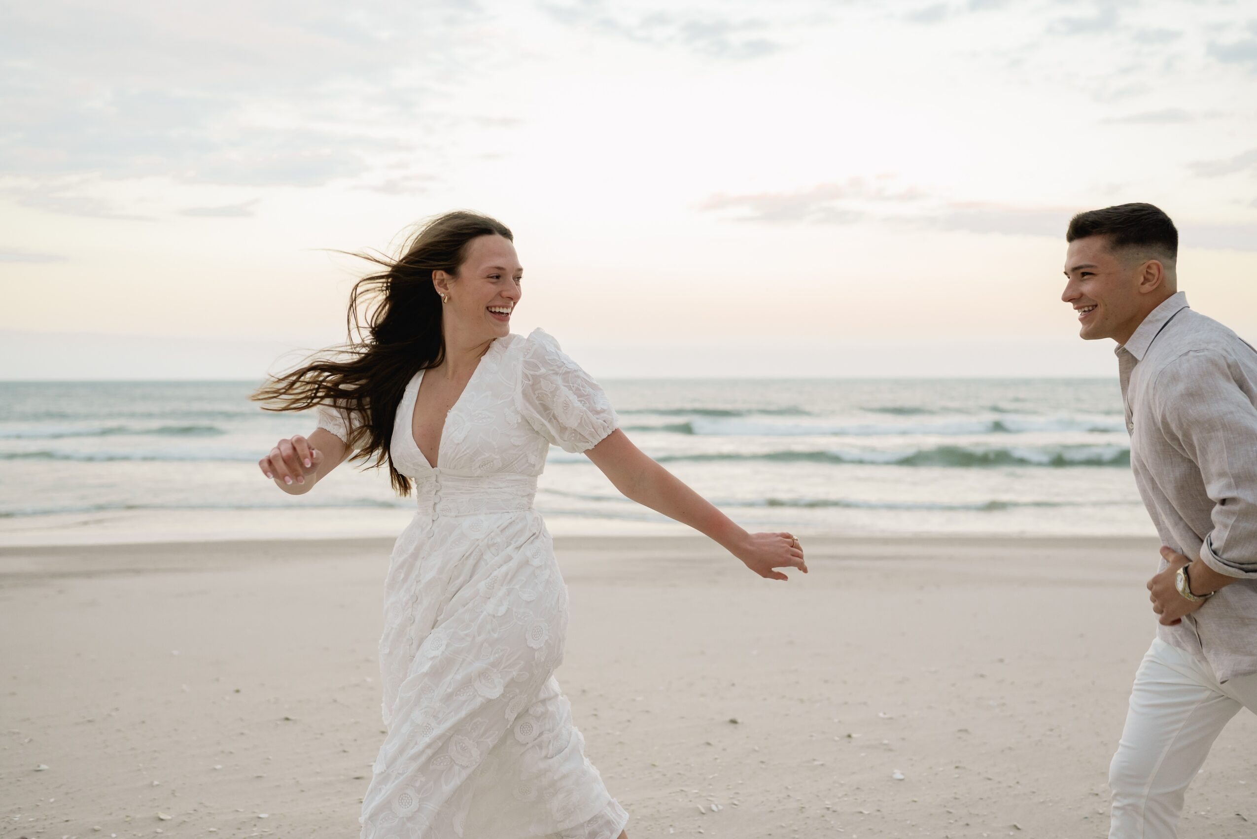 A woman in a white dress and a man in light clothing on a beach, smiling and holding hands, with ocean and sky in background.