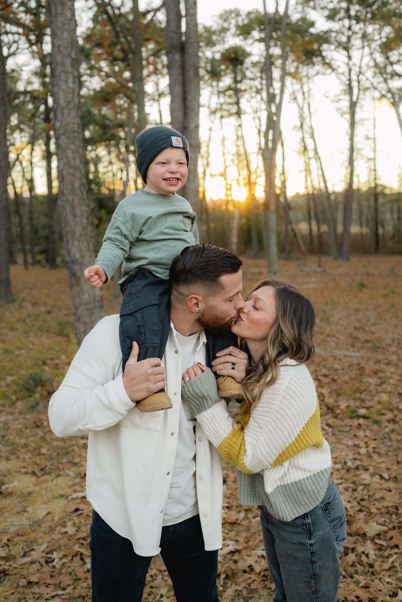 Family of three outdoors in autumn, child on father's shoulders, sunset in background, trees around, smiling faces.