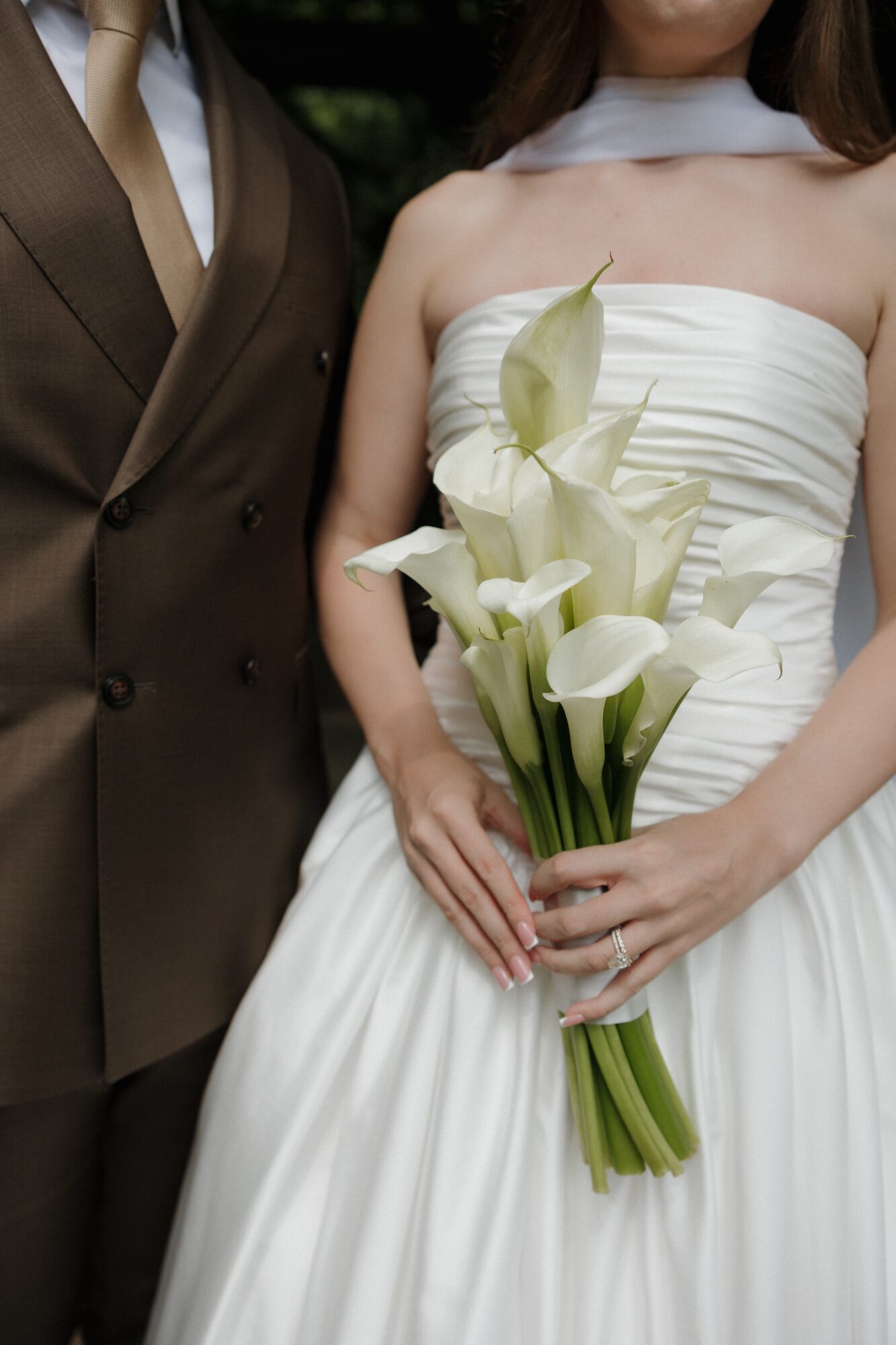 Bride in white strapless dress holding a bouquet of white calla lilies, standing next to a person in a brown suit.