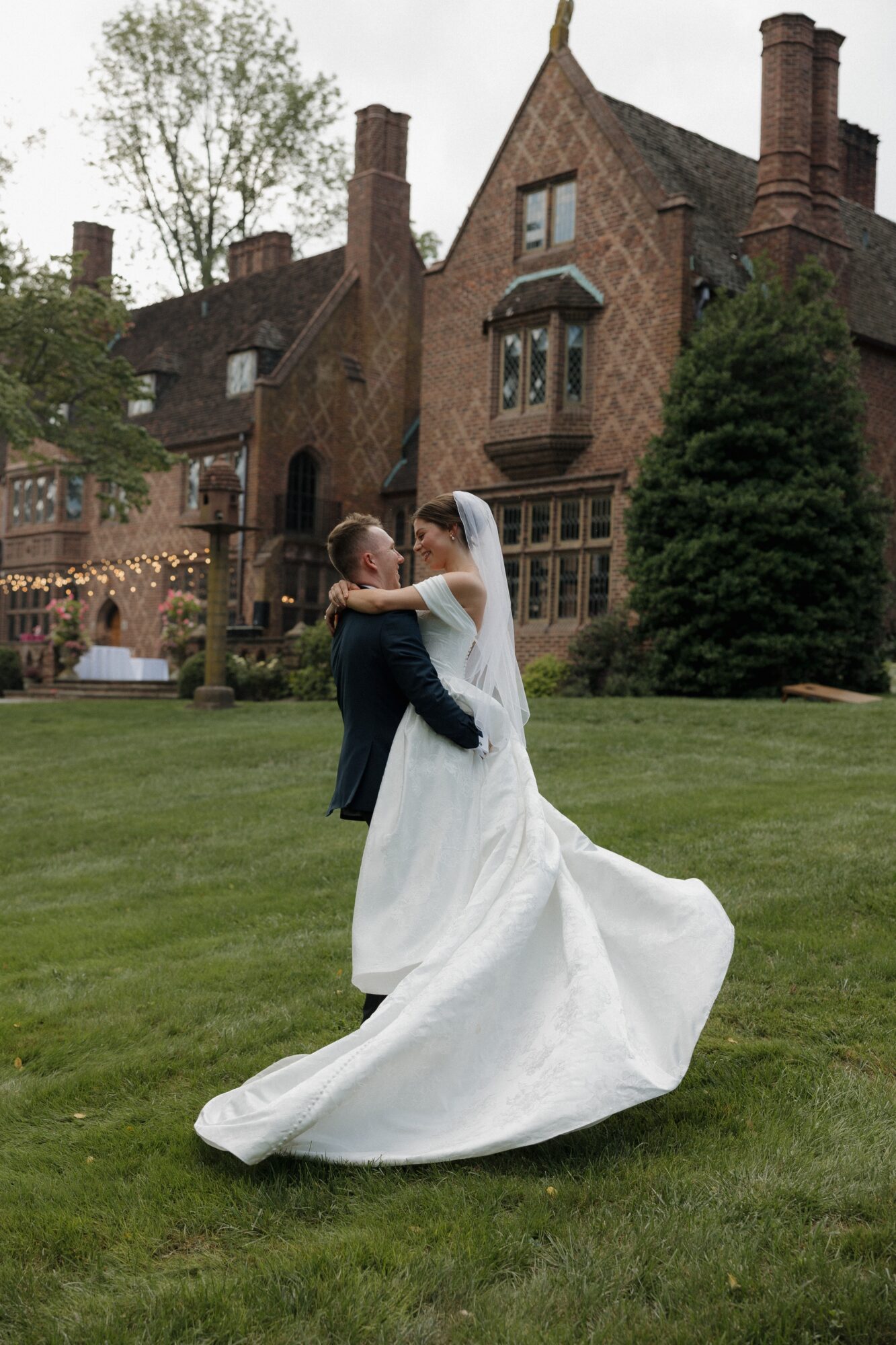 Bride and groom dancing on grass in front of a large historic brick house with trees and windows.