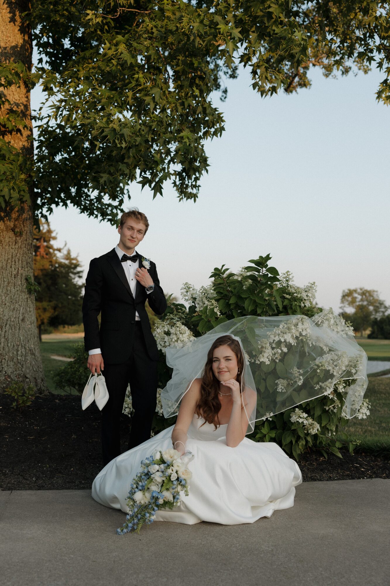 A bride in a white wedding dress and a groom in a black tuxedo stand outdoors near a tree and a bush, with a clear sky in the background.