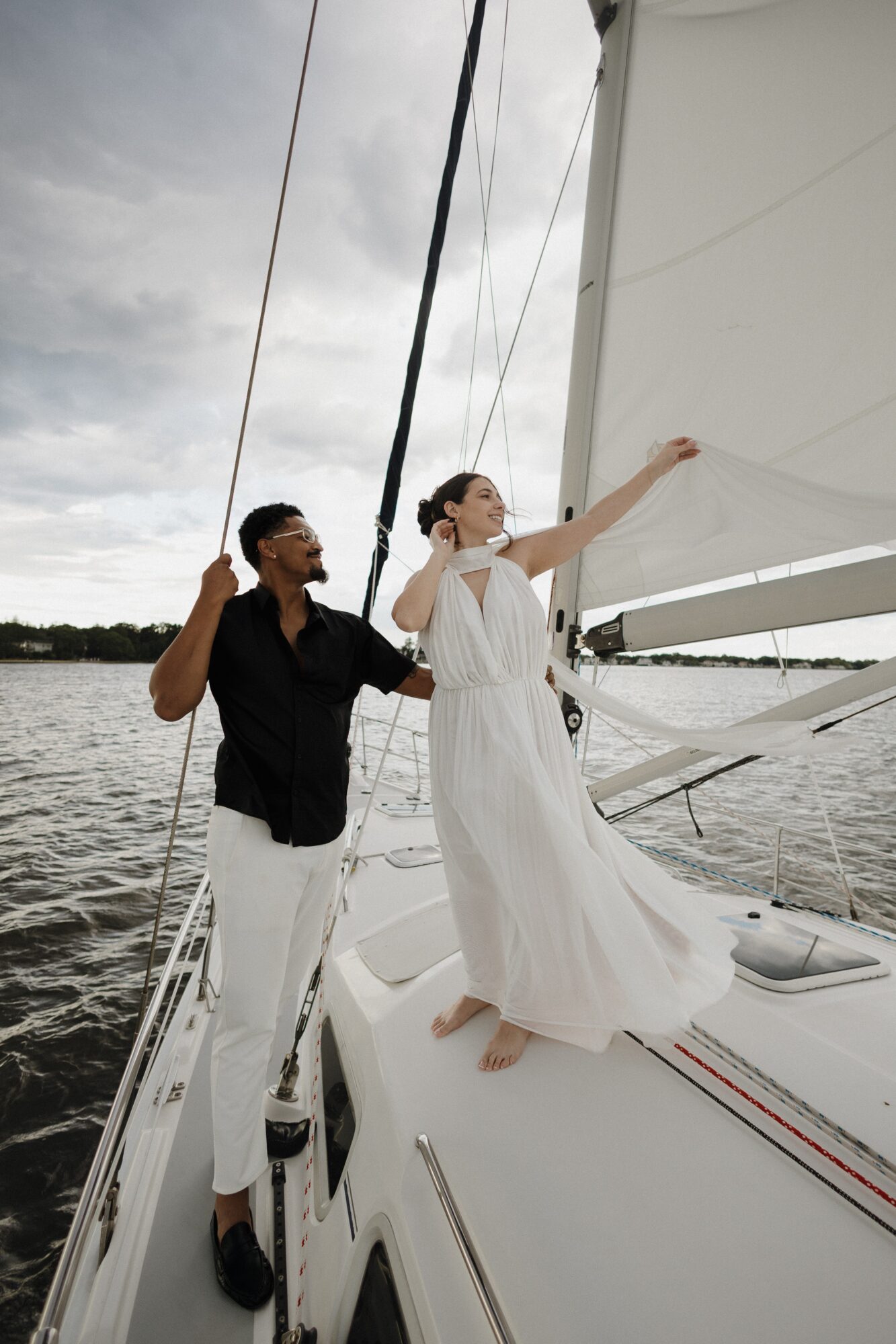 Two people on a sailboat, one adjusting the sail, the other standing barefoot, with water and cloudy sky in background.