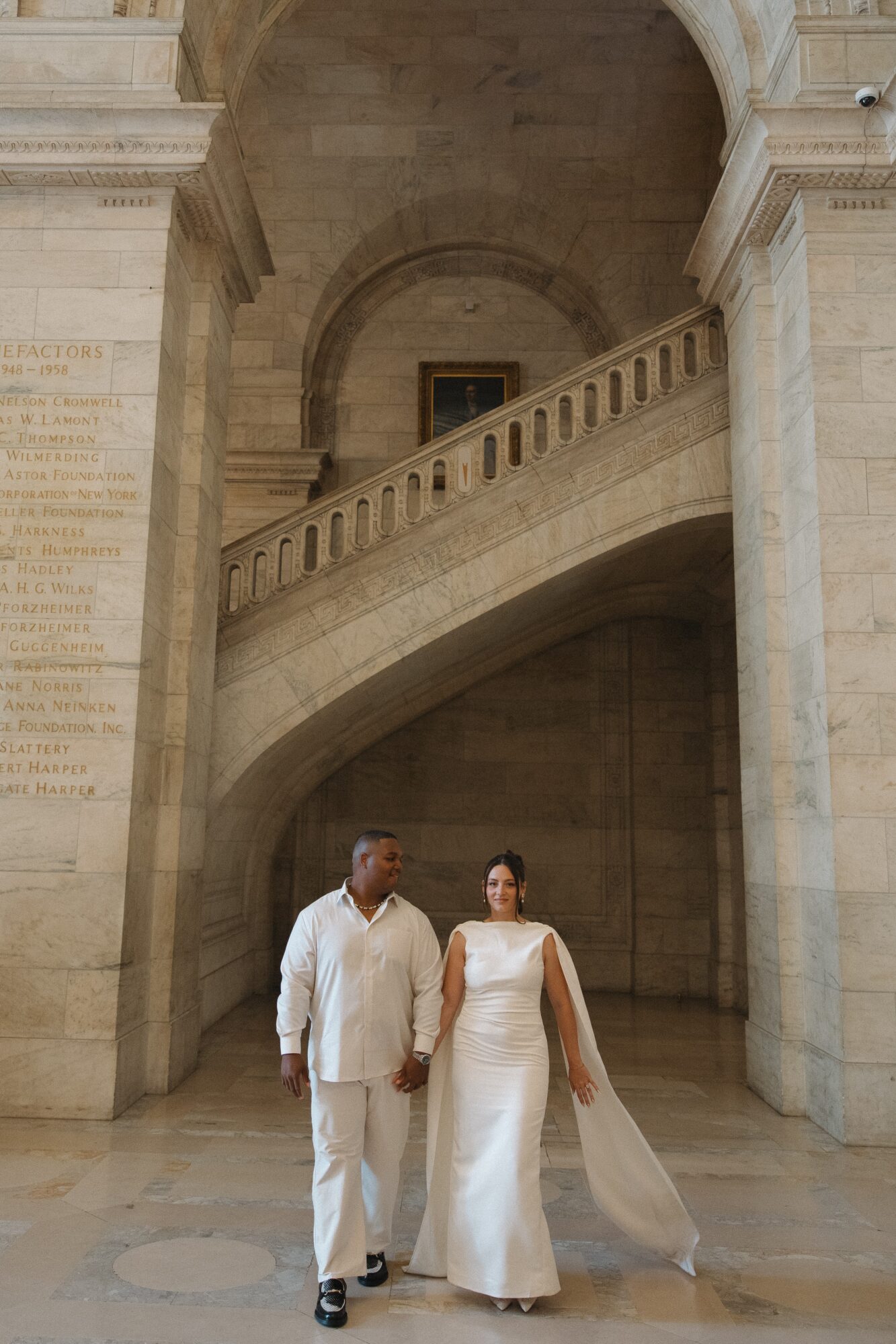 A couple in wedding attire walking inside a grand stone building with arches and columns.