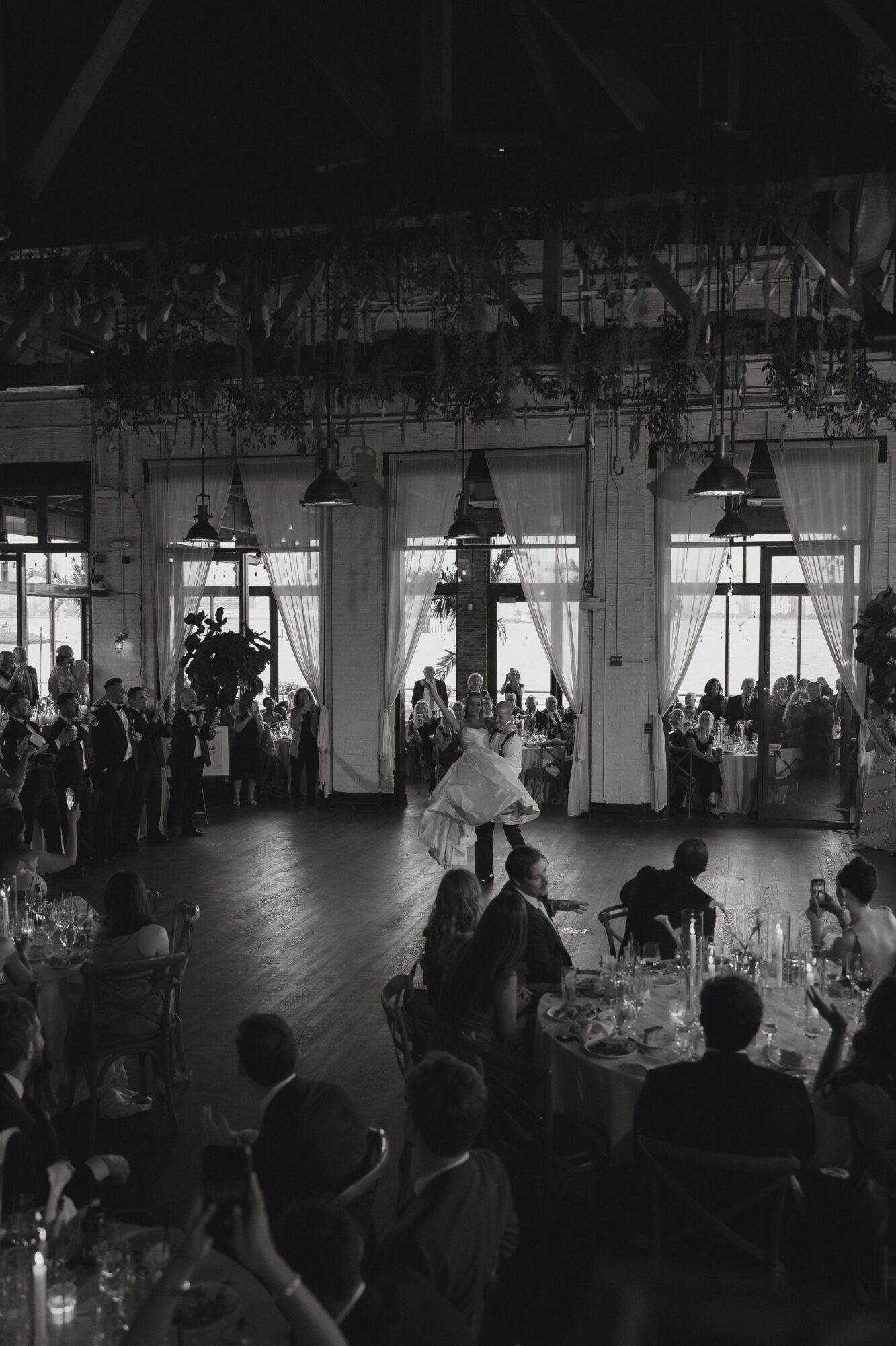 People dancing in a large indoor space with tables and large windows, some guests seated and others standing or dancing.