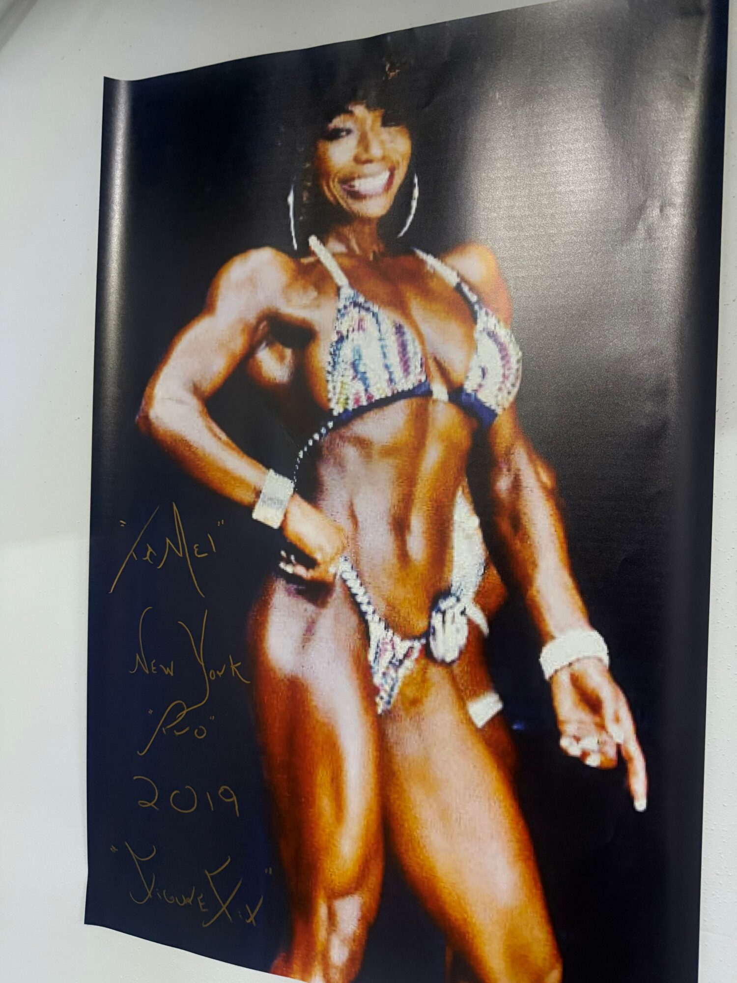 Bodybuilder in a competition pose, wearing a decorated bikini, pointing with one arm, on a dark background.