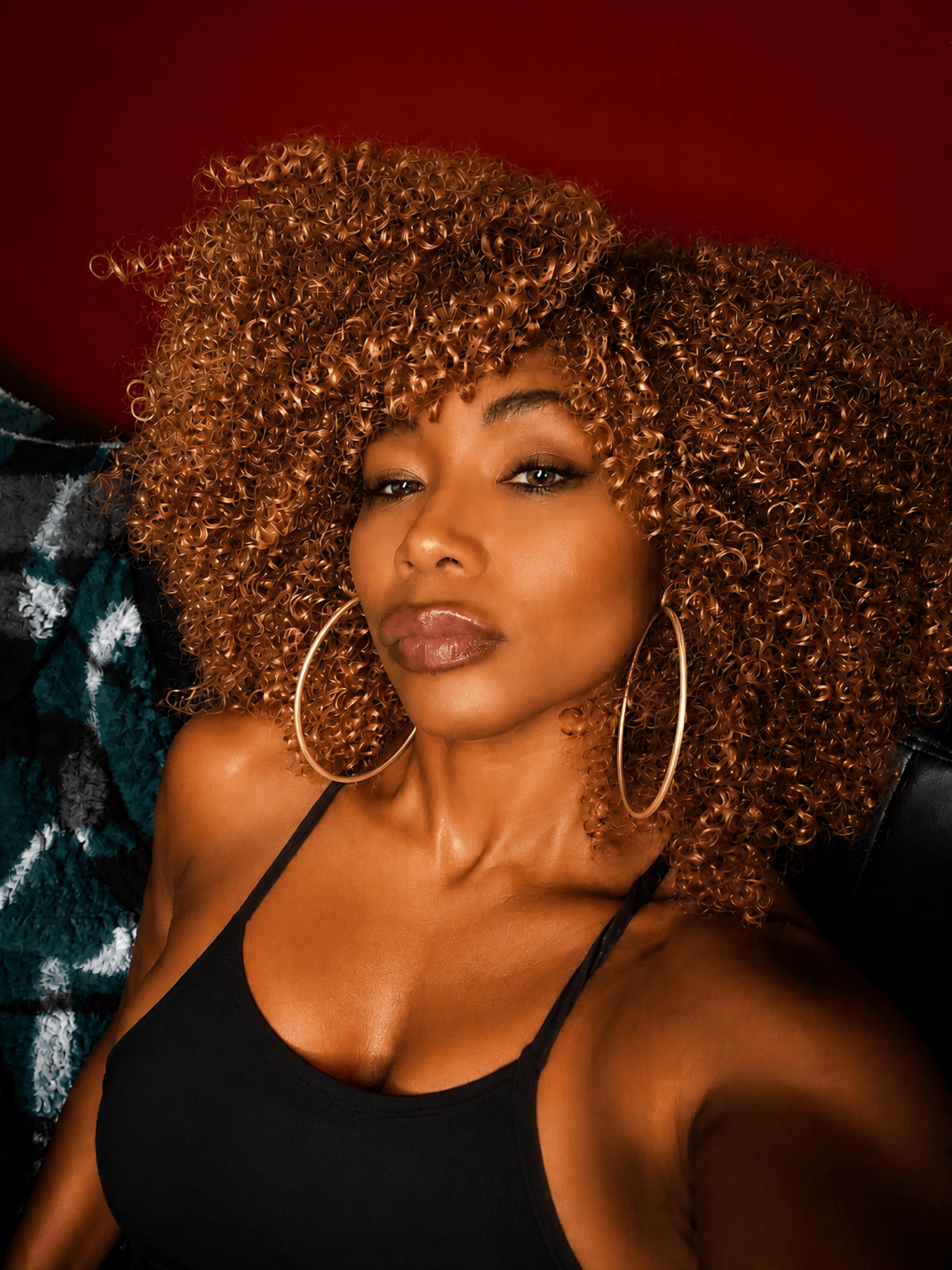 Woman with curly hair, large hoop earrings, wearing a black top, posing indoors with a red background.