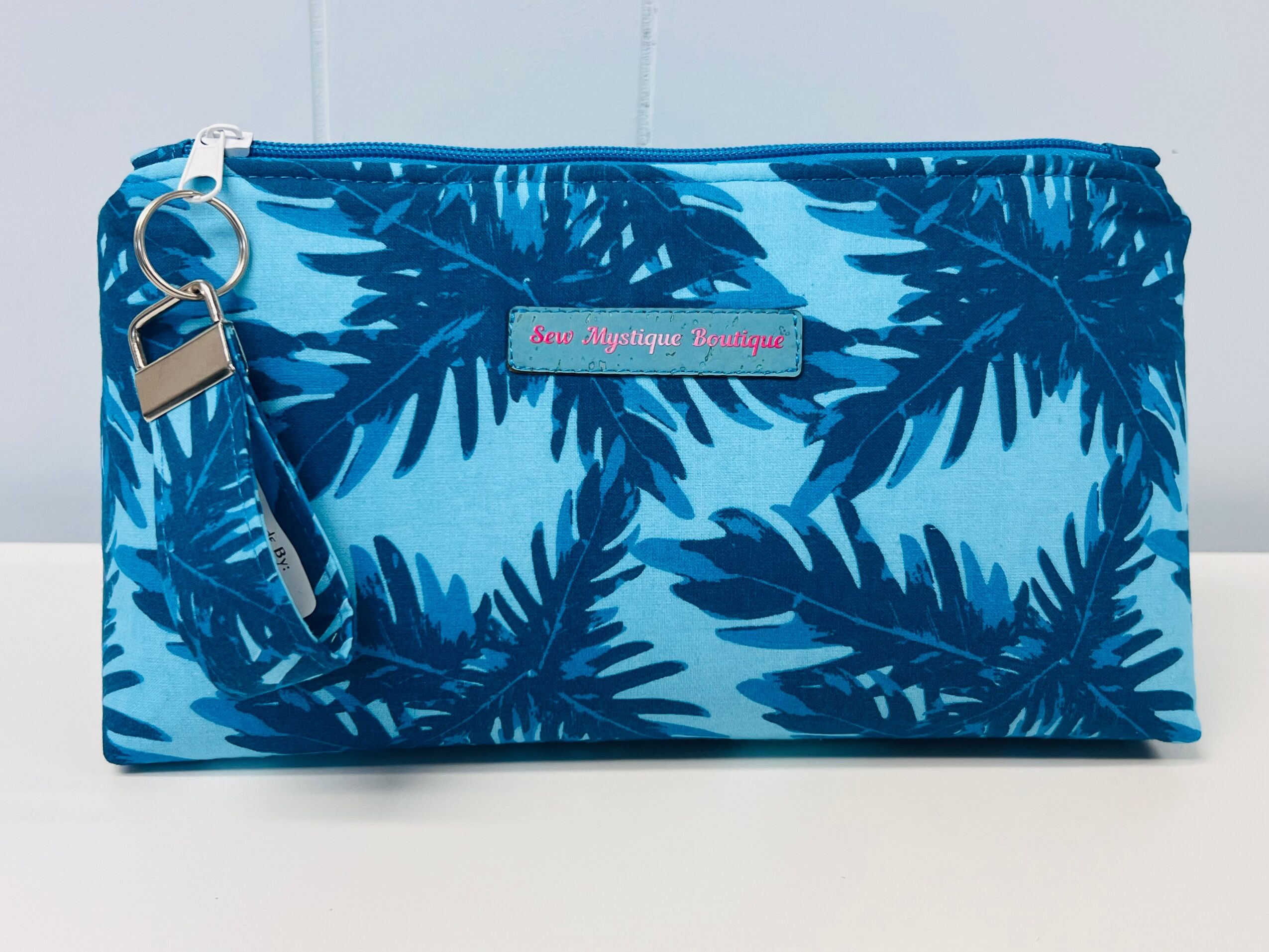 Blue tropical leaf-patterned zippered pouch with a keyring attachment, placed on a white surface.