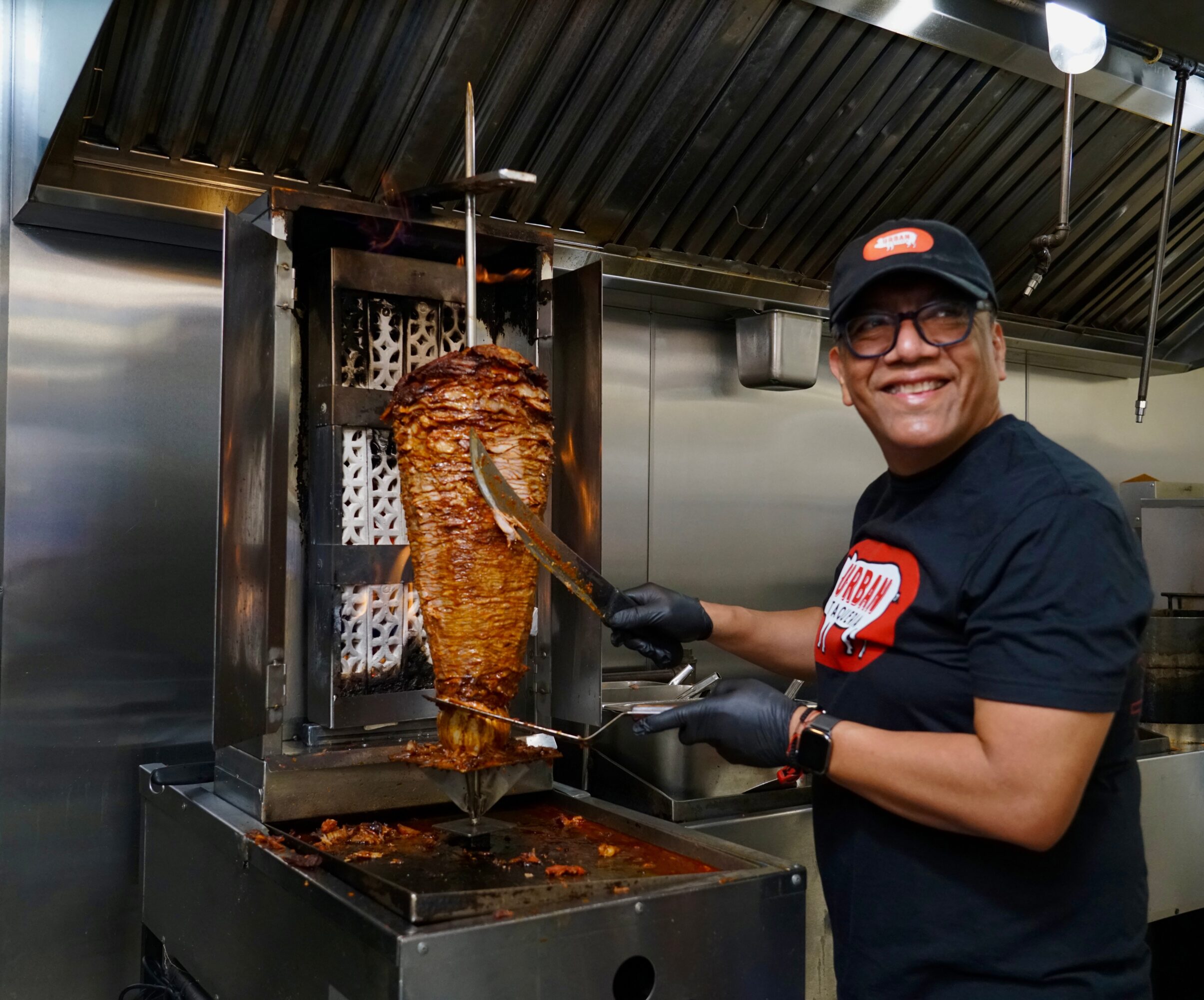 Person wearing glasses and a cap, smiling, prepares meat on a vertical rotisserie in a kitchen.