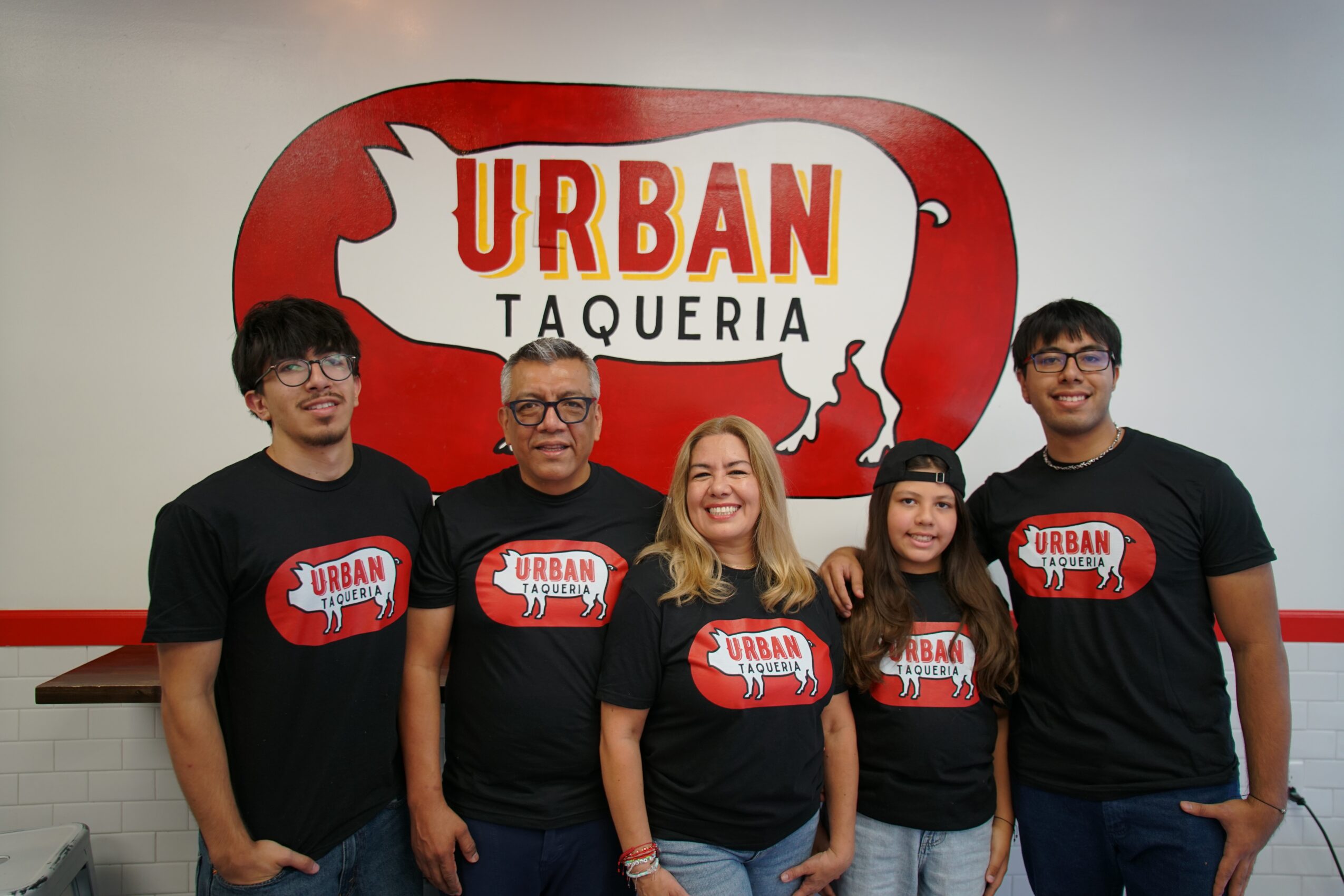 Group of five people wearing matching black T-shirts with a red and white logo, standing in front of a sign that says 'URBAN TAQUERIA'.