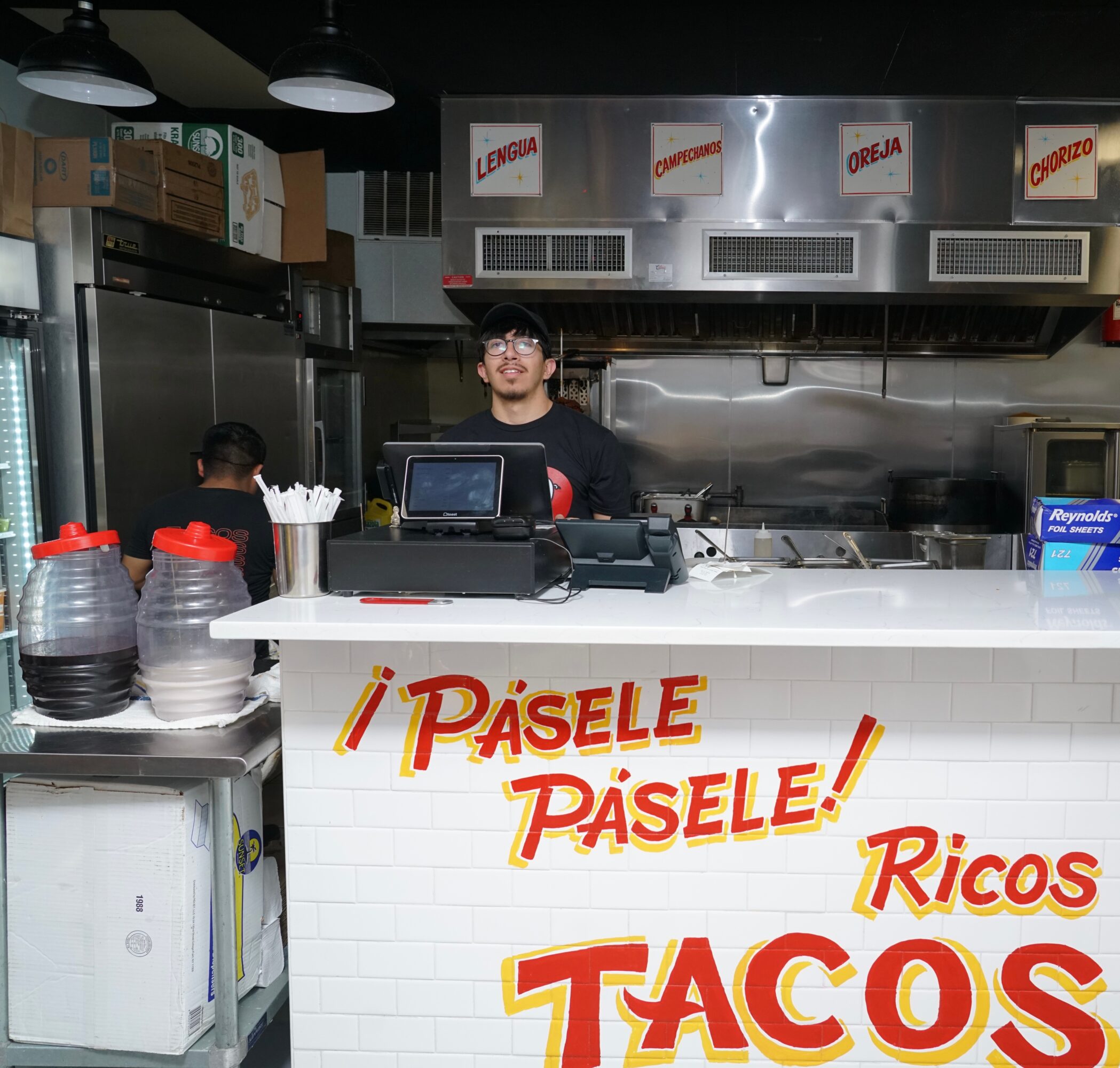 Person standing behind a taco stand counter with signs and kitchen equipment, smiling at the camera.