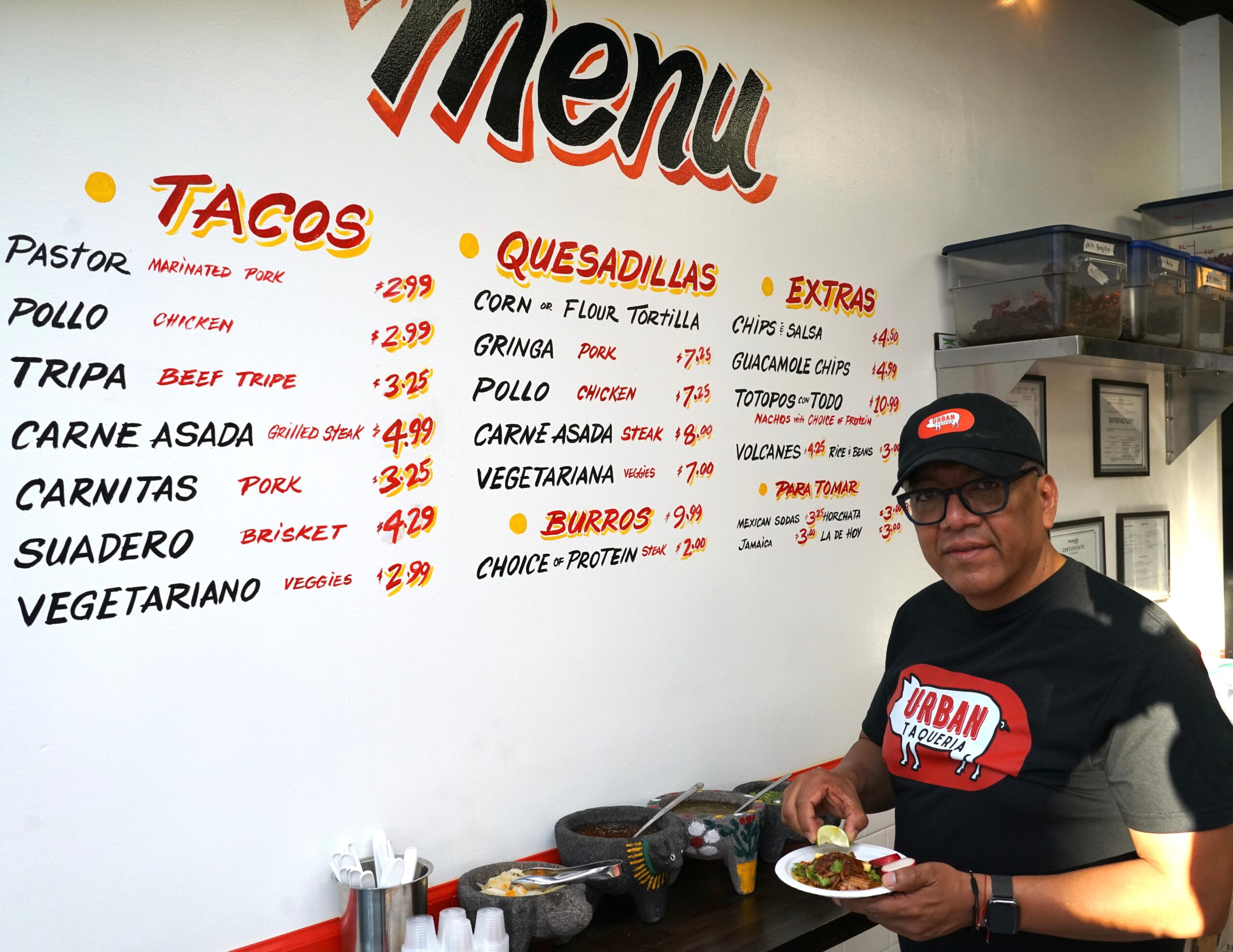 Person wearing glasses and a black shirt with a red and white logo, holding a plate of food in front of a menu board.