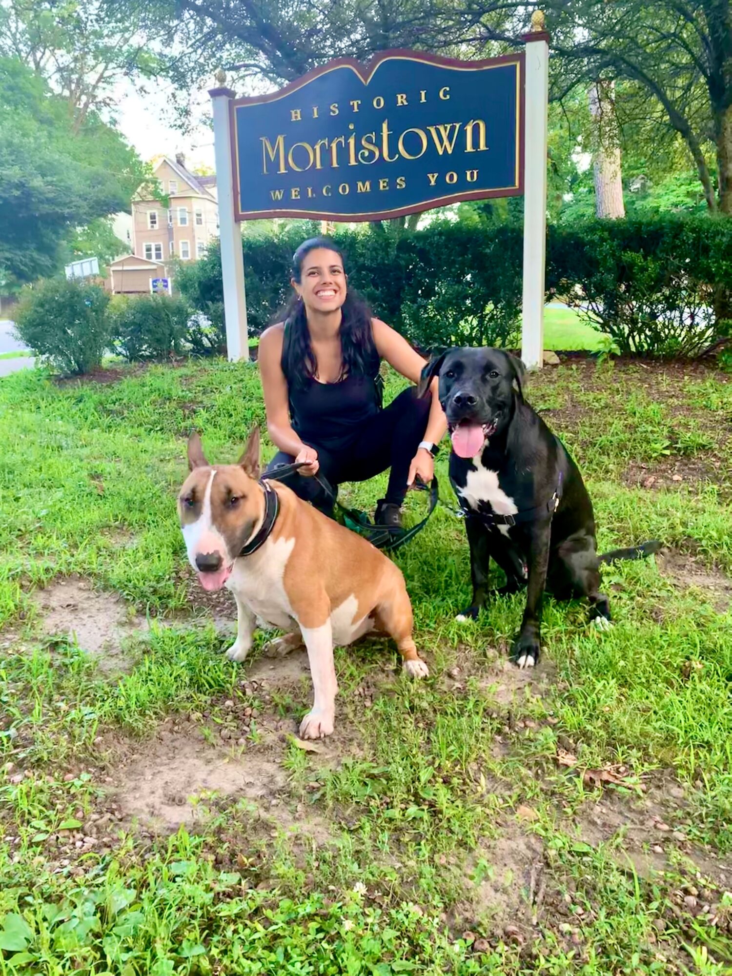 Woman with two dogs in front of Morristown sign, smiling outdoors on grass and trees.