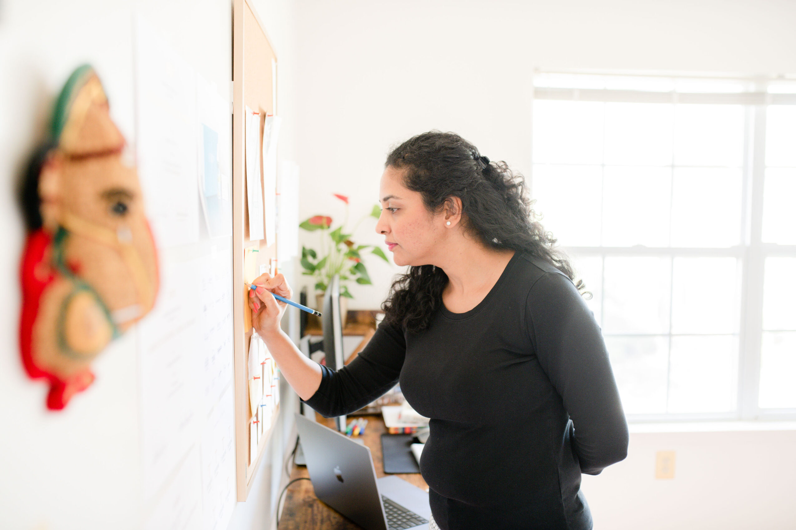Woman with dark curly hair writing on a whiteboard in a bright room with large windows, a laptop nearby.
