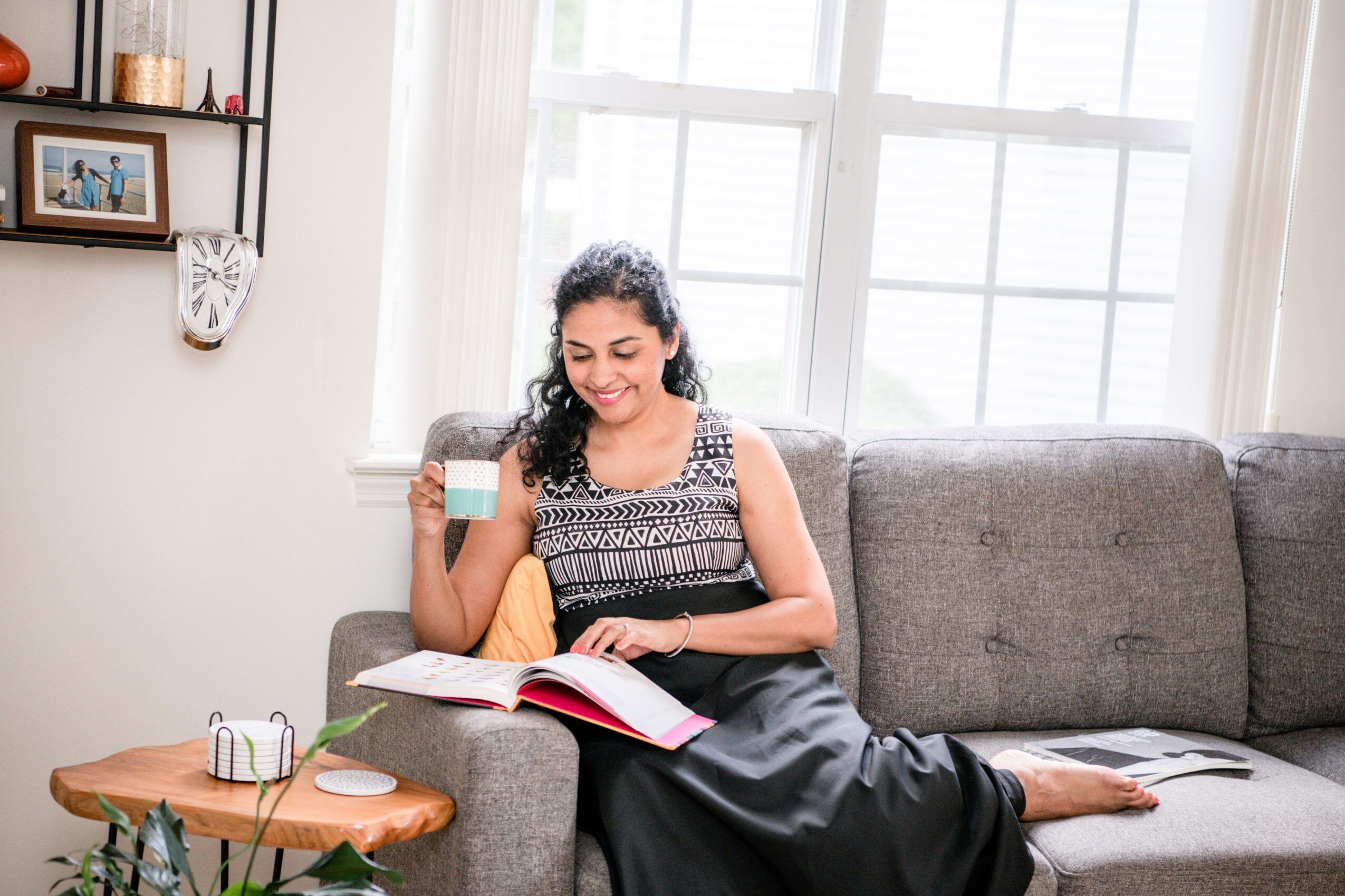 Woman sitting on a sofa reading a book, holding a mug, in a bright room with large windows
