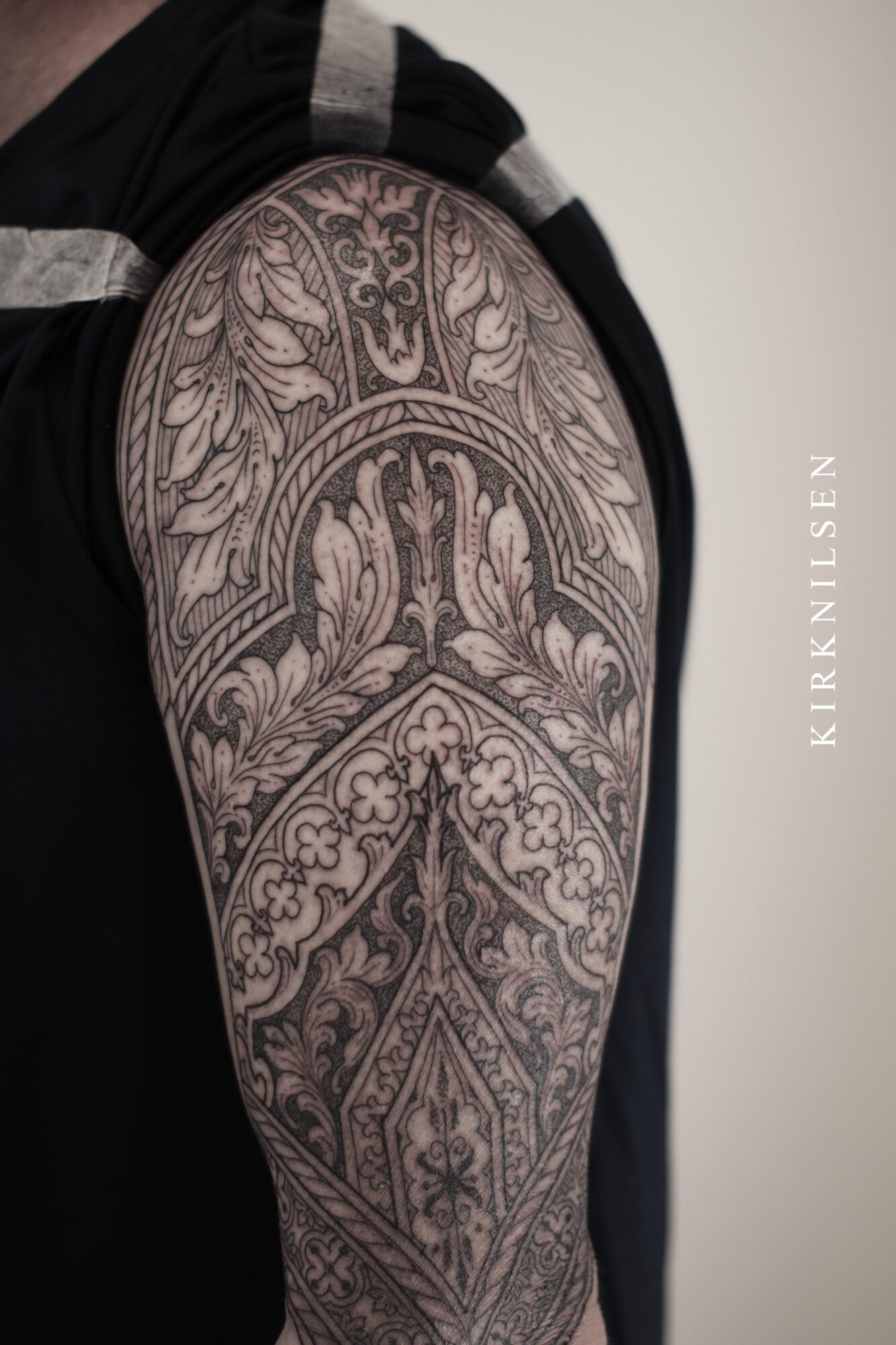 Close-up of a person's shoulder with detailed black and gray tattoo featuring floral and ornamental patterns.