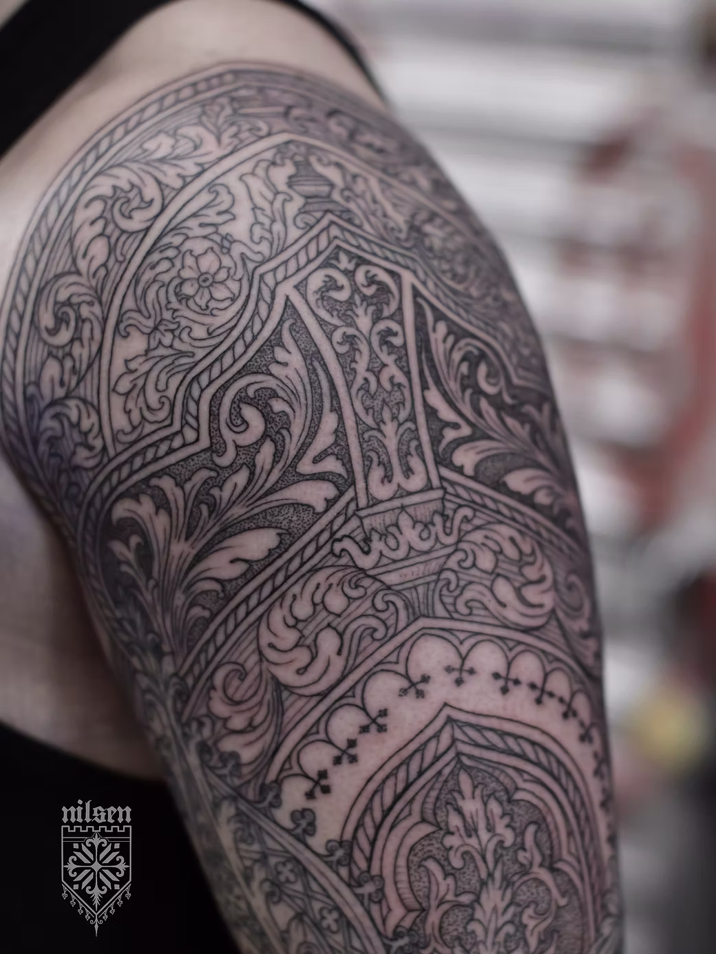 Close-up of a detailed black and gray tattoo on a person's shoulder and upper arm, featuring intricate patterns and designs.