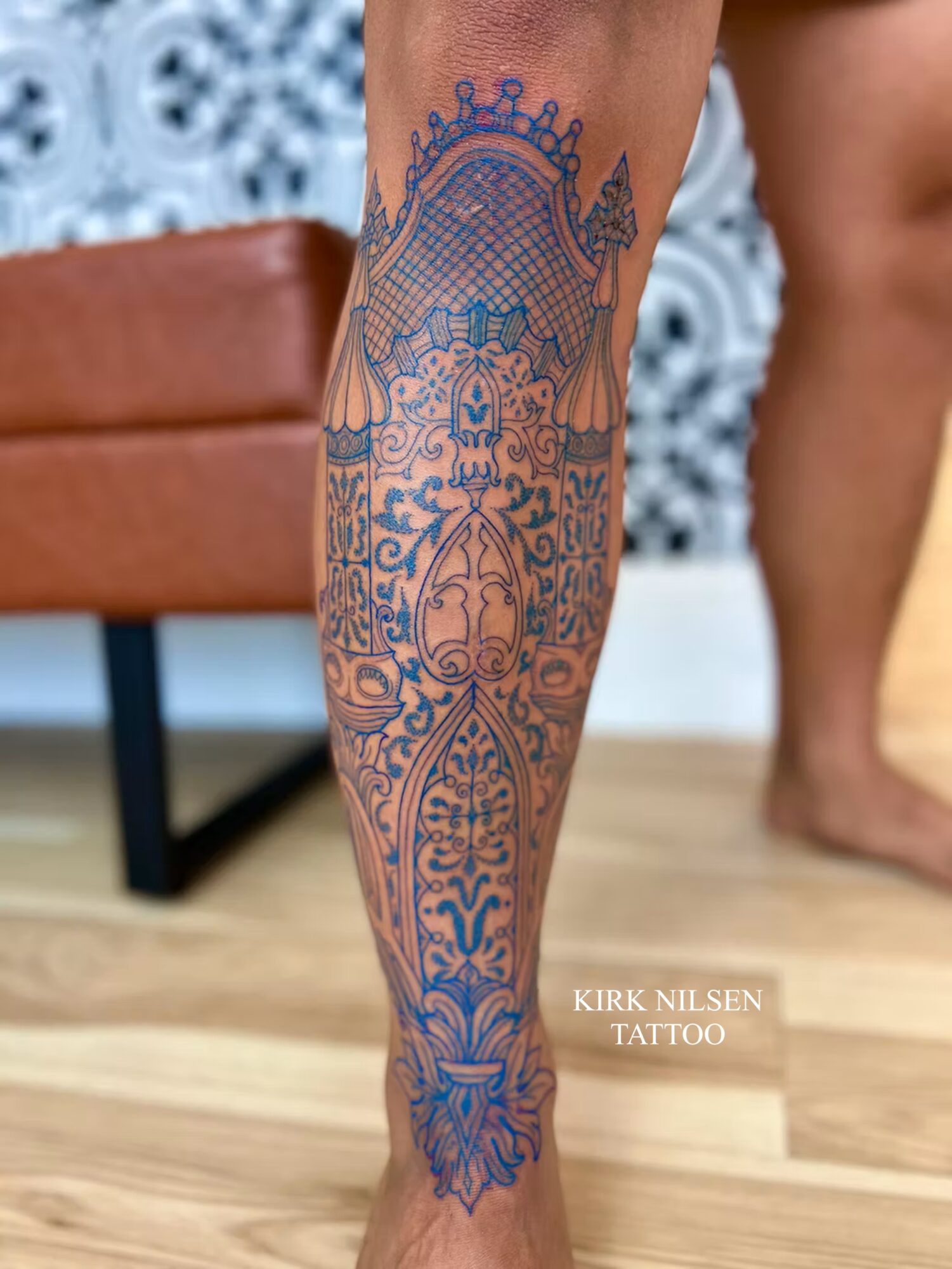Tattooed leg with intricate blue design extending from knee to ankle, with a background of a wooden floor and furniture.