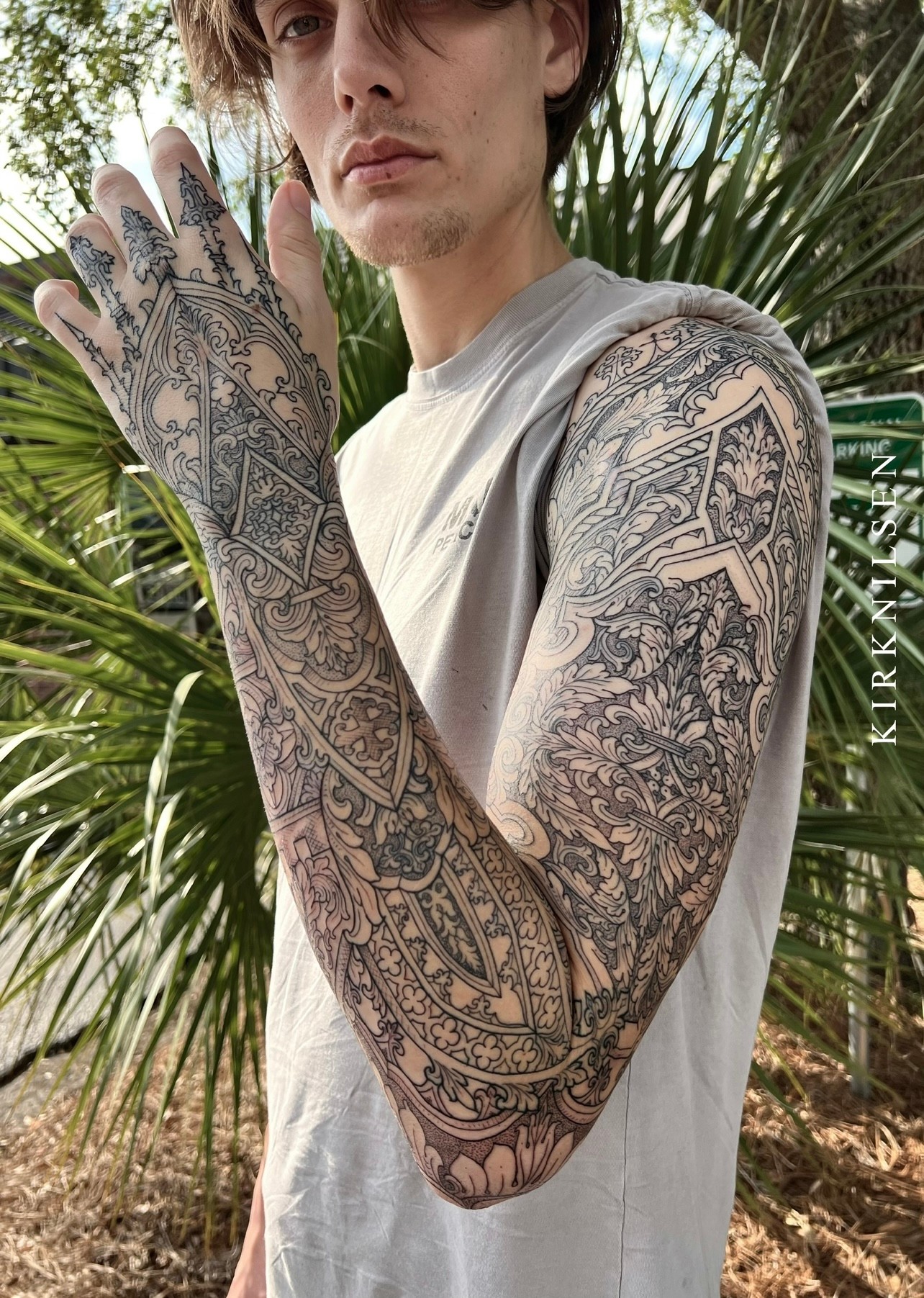 Person with tattoos on arm and hand, outdoors with green plants in background, wearing a sleeveless shirt.
