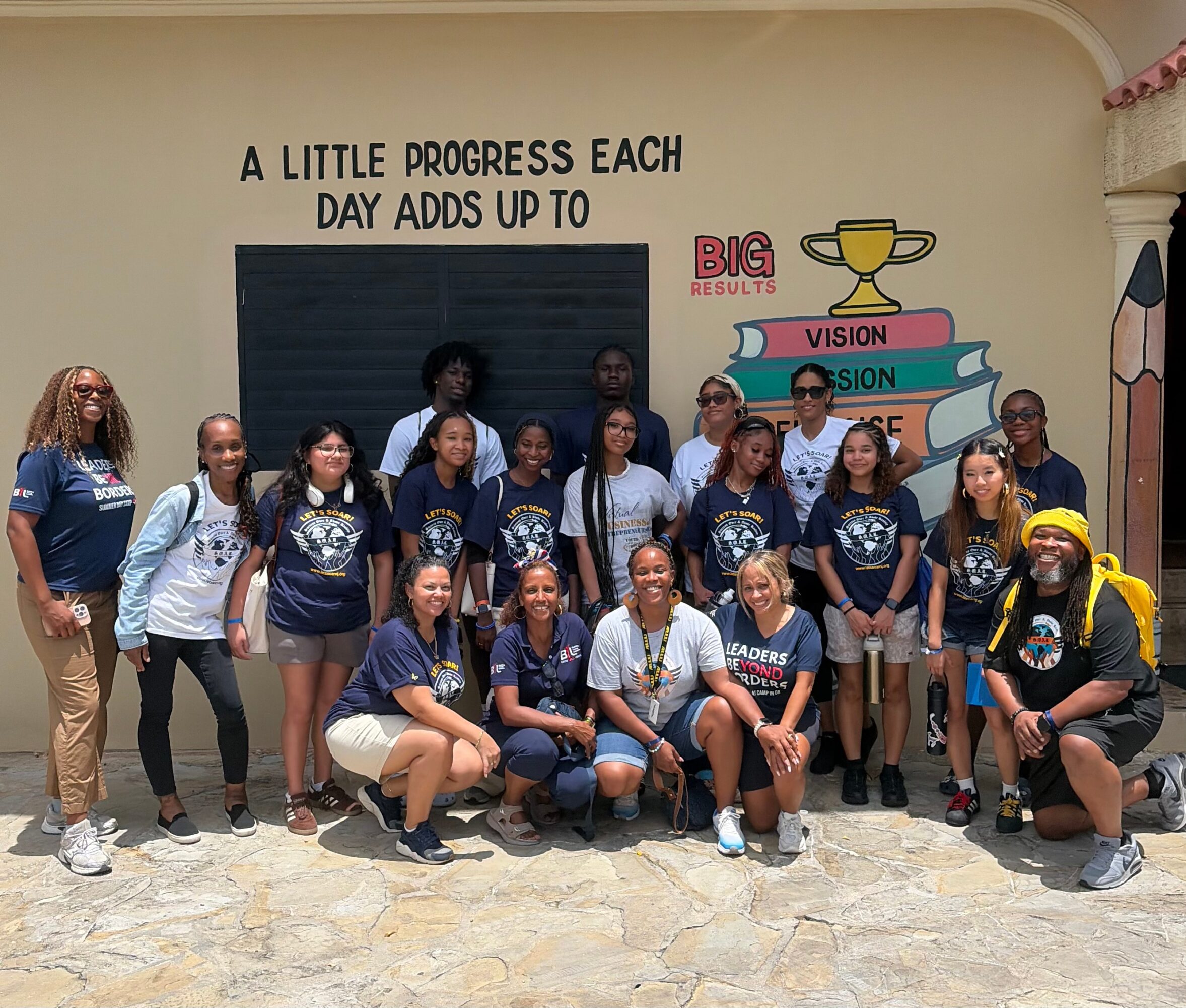 Group of young girls and women standing outdoors in front of a wall with motivational text and illustrations, some smiling.