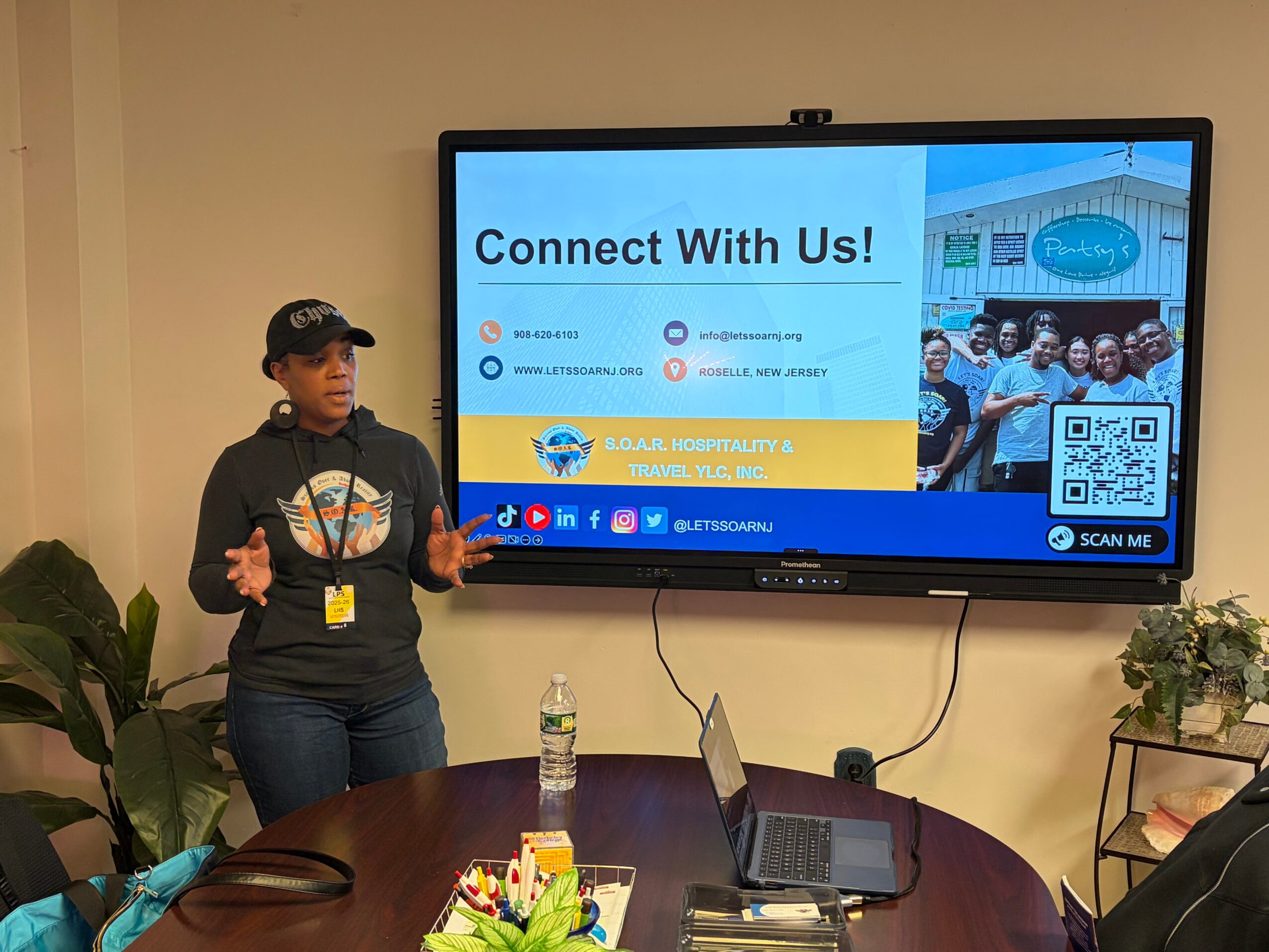 Woman presenting in front of a screen displaying a community connection slide, with a table and plants nearby.