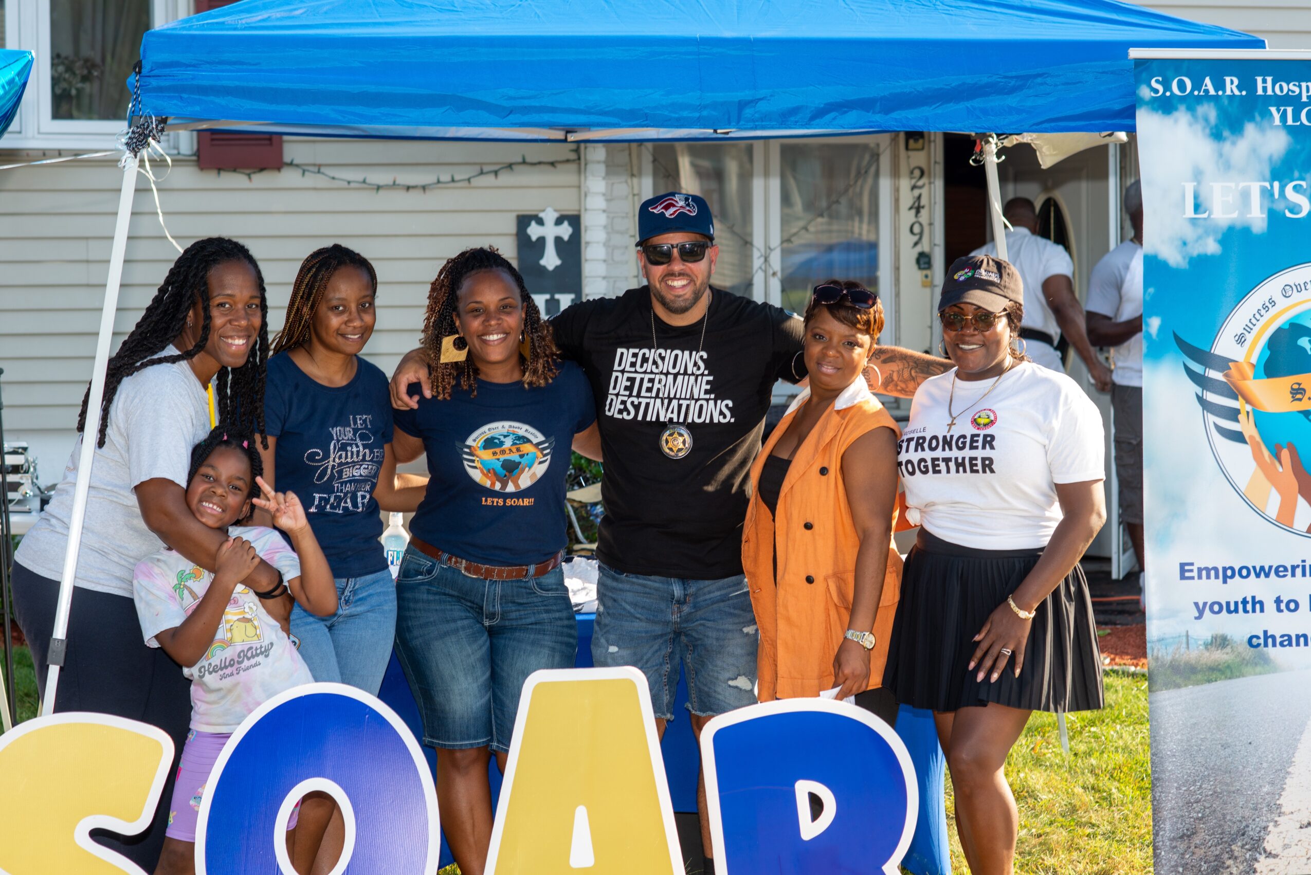 Group of seven people standing outdoors under a blue canopy, smiling, with a banner on the right side, in front of a building.
